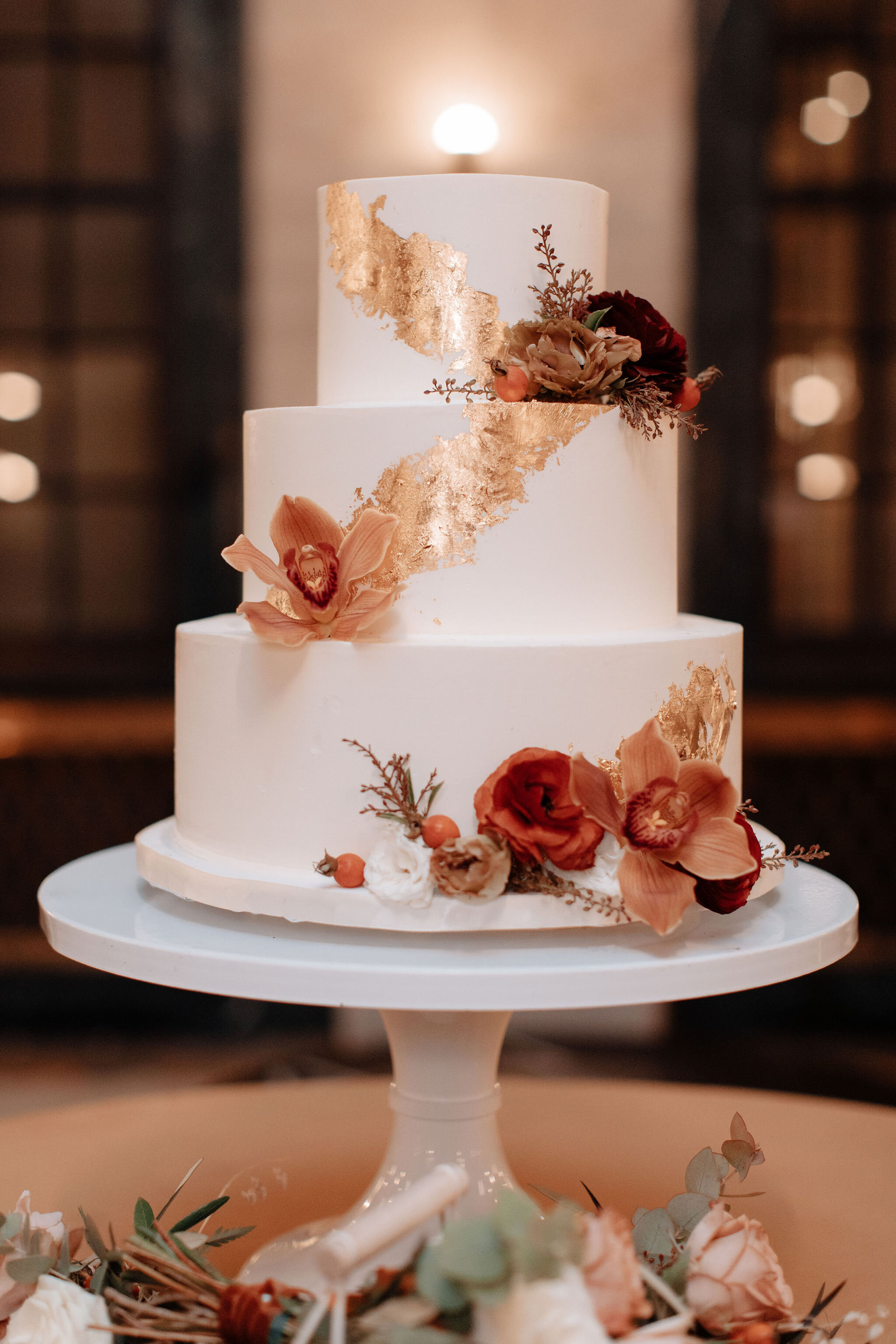 Gold leaf accents for the cake with rust orange orchids, dusty red ranunculus, and muted textures.