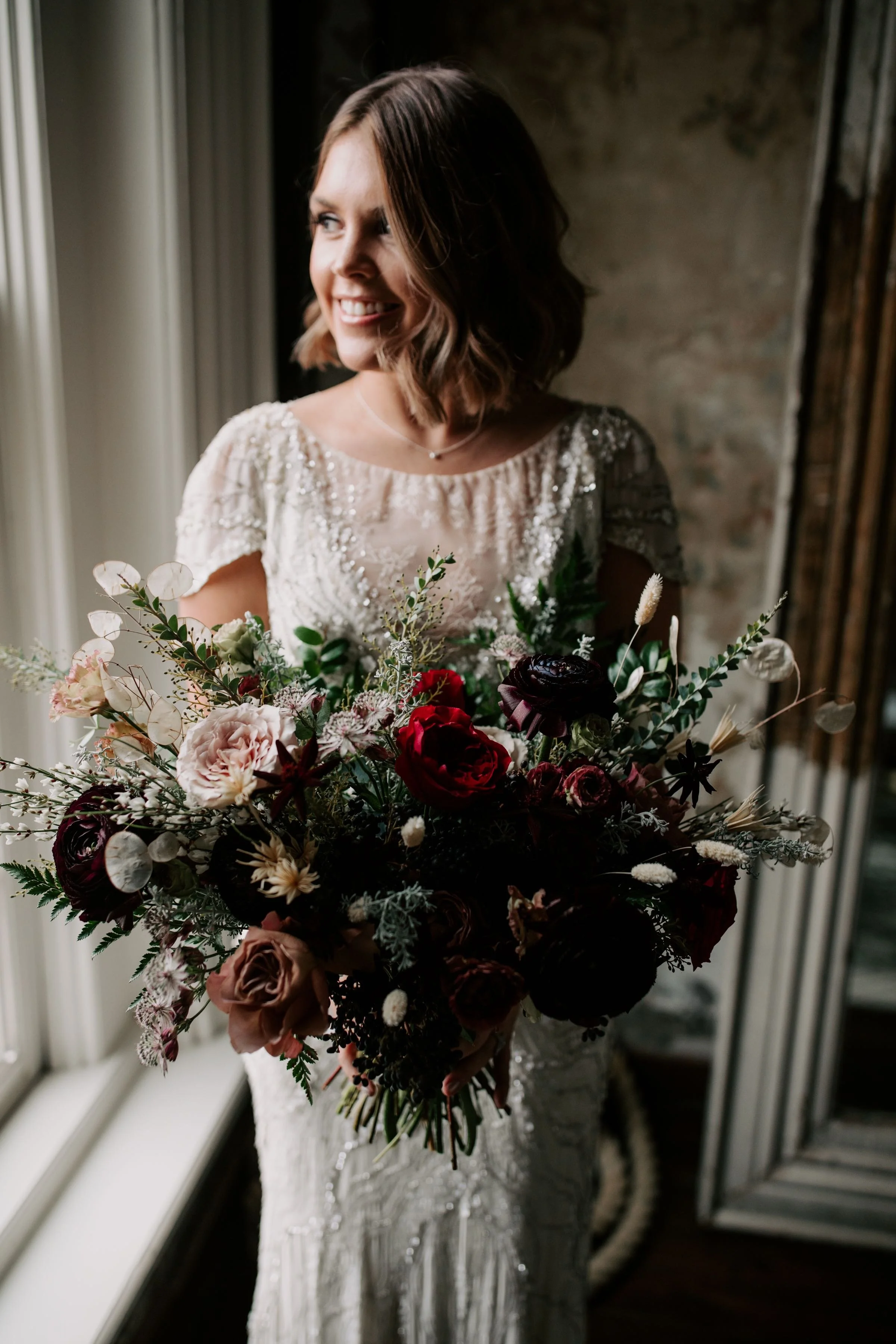 Loose, whimsical winter bridal bouquet with eggplant, blush, mauve, and burgundy garden roses and ranunculus, dried flowers, and natural, untamed greenery. Nashville wedding floral design at the Cordelle.