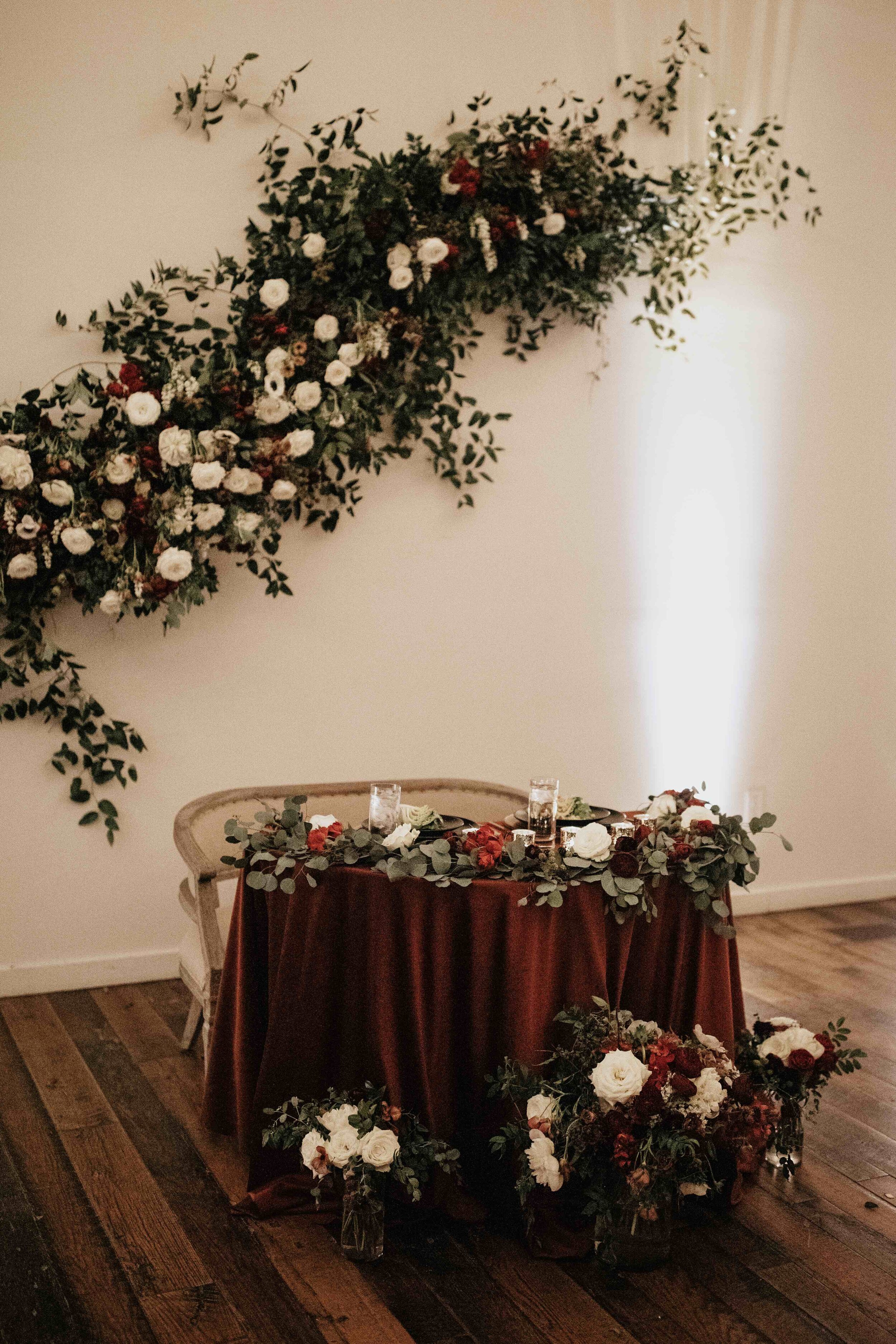 Asymmetrical floral installation growing up the white wall of the Cordelle with natural, untamed greenery, burgundy and white garden roses and ranunculus, and organic textures. Nashville wedding florist.