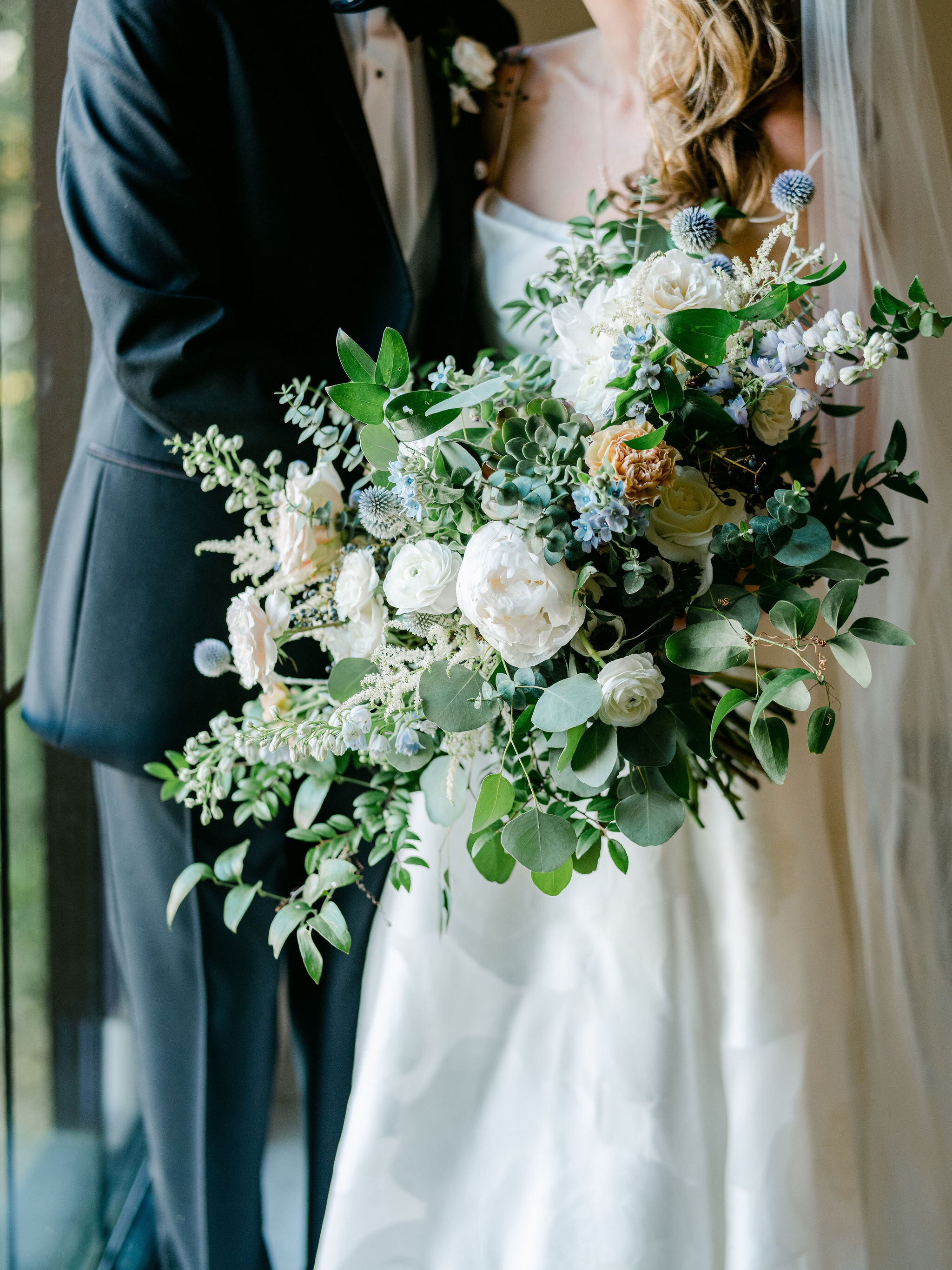 A lush and sprawling bridal bouquet filled to the brim. Blush and white garden roses, white peonies, blue thistle, delphinium, blue tweedia, white astilbe, white ranunculus, baby and silver dollar eucalyptus, huckleberry, and smilax vine are just so…