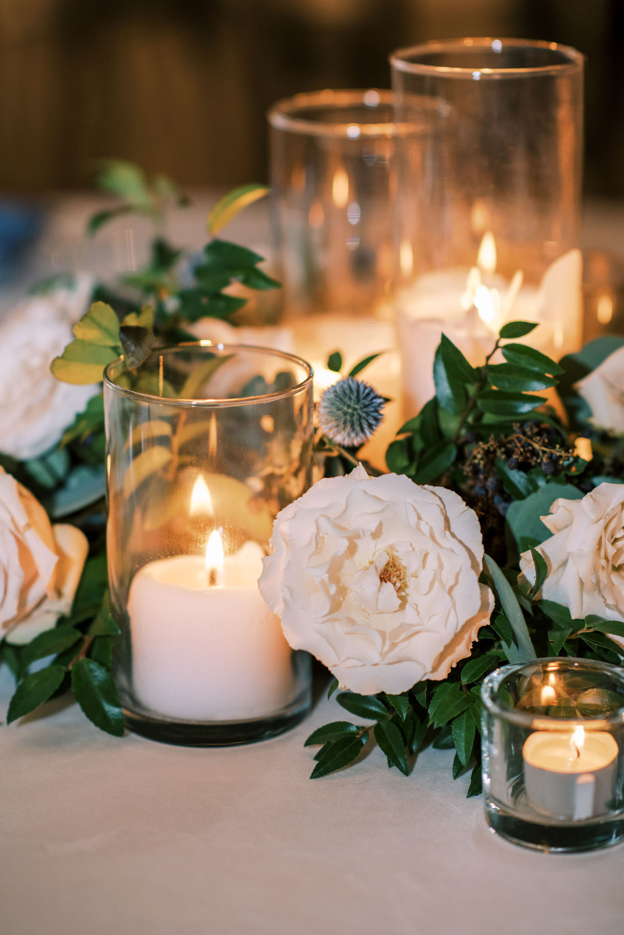 This wedding featured lush table garlands filled with blooms of blush and white garden roses, blue globe thistle, privet berry, seeded eucalyptus and lush greenery. Twinkly pillar candles and votives were scattered throughout the garland. Designed b…