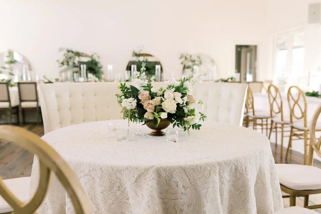 This twinkling reception is filled with taupe tapers in collected brass candlesticks. Centerpieces filled with delicate blooms of quicksand roses, white ice roses, majolica spray roses, fluffy ranunculus, white stock, delicate sweet pea and natural …