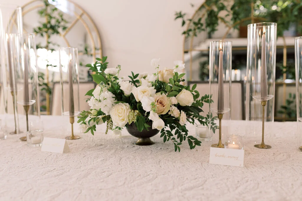 This twinkling reception is filled with taupe tapers in collected brass candlesticks. Centerpieces filled with delicate blooms of quicksand roses, white ice roses, majolica spray roses, fluffy ranunculus, white stock, delicate sweet pea and natural …