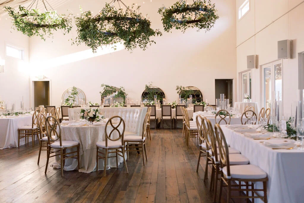 This twinkling reception is filled with taupe tapers in collected brass candlesticks. Wild green chandeliers float over centerpieces filled with delicate blooms of quicksand roses, white ice roses, majolica spray roses, fluffy ranunculus, white stoc…