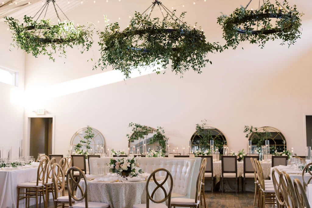 This twinkling reception is filled with taupe tapers in collected brass candlesticks. Wild green chandeliers float over centerpieces filled with delicate blooms of quicksand roses, white ice roses, majolica spray roses, fluffy ranunculus, white stoc…