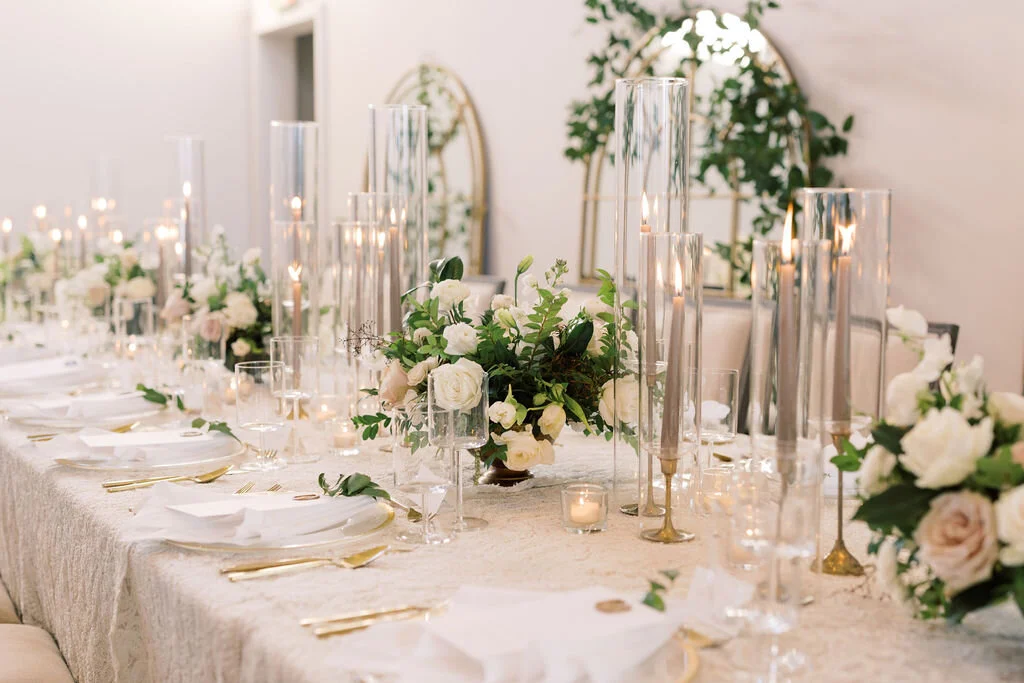 This twinkling reception is filled with taupe tapers in collected brass candlesticks. Centerpieces filled with delicate blooms of quicksand roses, white ice roses, majolica spray roses, fluffy ranunculus, white stock, delicate sweet pea and natural …