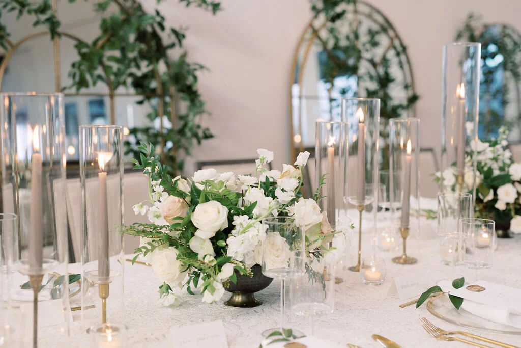 This twinkling reception is filled with taupe tapers in collected brass candlesticks. Centerpieces filled with delicate blooms of quicksand roses, white ice roses, majolica spray roses, fluffy ranunculus, white stock, delicate sweet pea and natural …