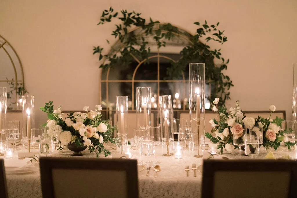 This twinkling reception is filled with taupe tapers in collected brass candlesticks. Centerpieces filled with delicate blooms of quicksand roses, white ice roses, majolica spray roses, fluffy ranunculus, white stock, delicate sweet pea and natural …