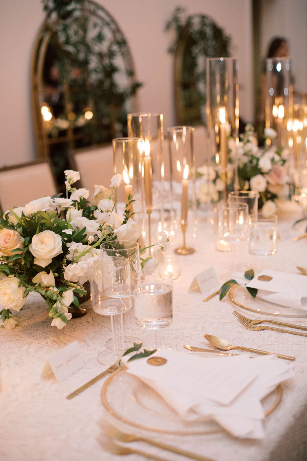 This twinkling reception is filled with taupe tapers in collected brass candlesticks. Centerpieces filled with delicate blooms of quicksand roses, white ice roses, majolica spray roses, fluffy ranunculus, white stock, delicate sweet pea and natural …