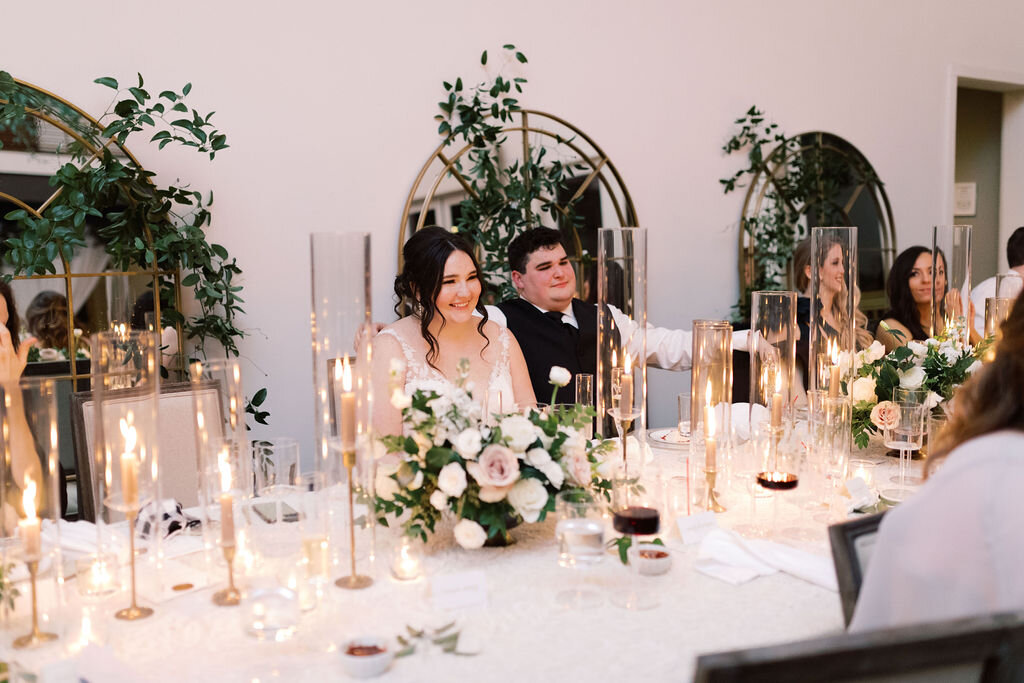 This twinkling reception is filled with taupe tapers in collected brass candlesticks. Centerpieces filled with delicate blooms of quicksand roses, white ice roses, majolica spray roses, fluffy ranunculus, white stock, delicate sweet pea and natural …