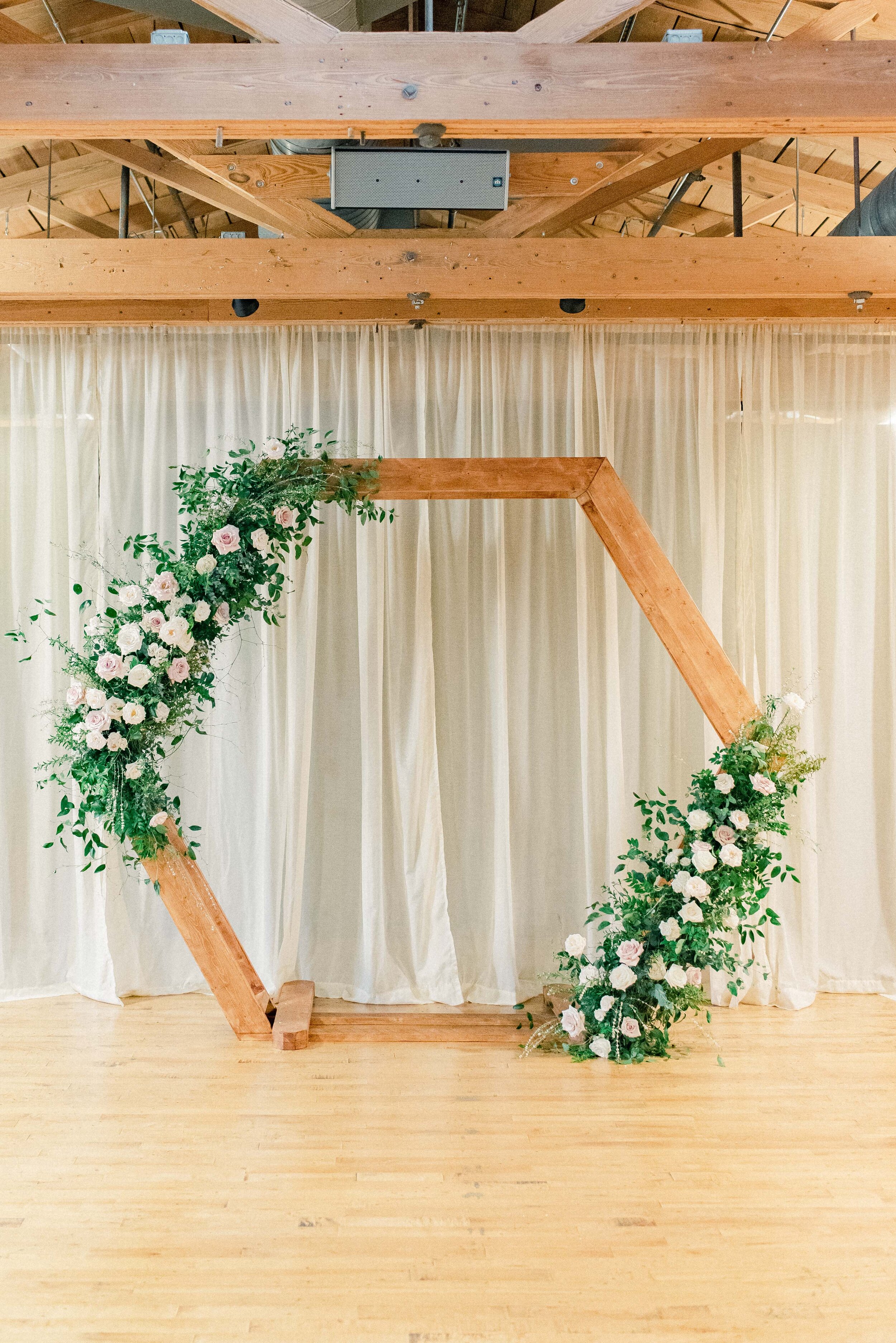 A hexagon arch makes the perfect ceremony backdrop. Blooms used here are blush quicksand roses, cream garden roses, white ranunculus, spray roses, and lush greenery. Designed by florist Rosemary & Finch in Nashville, TN.