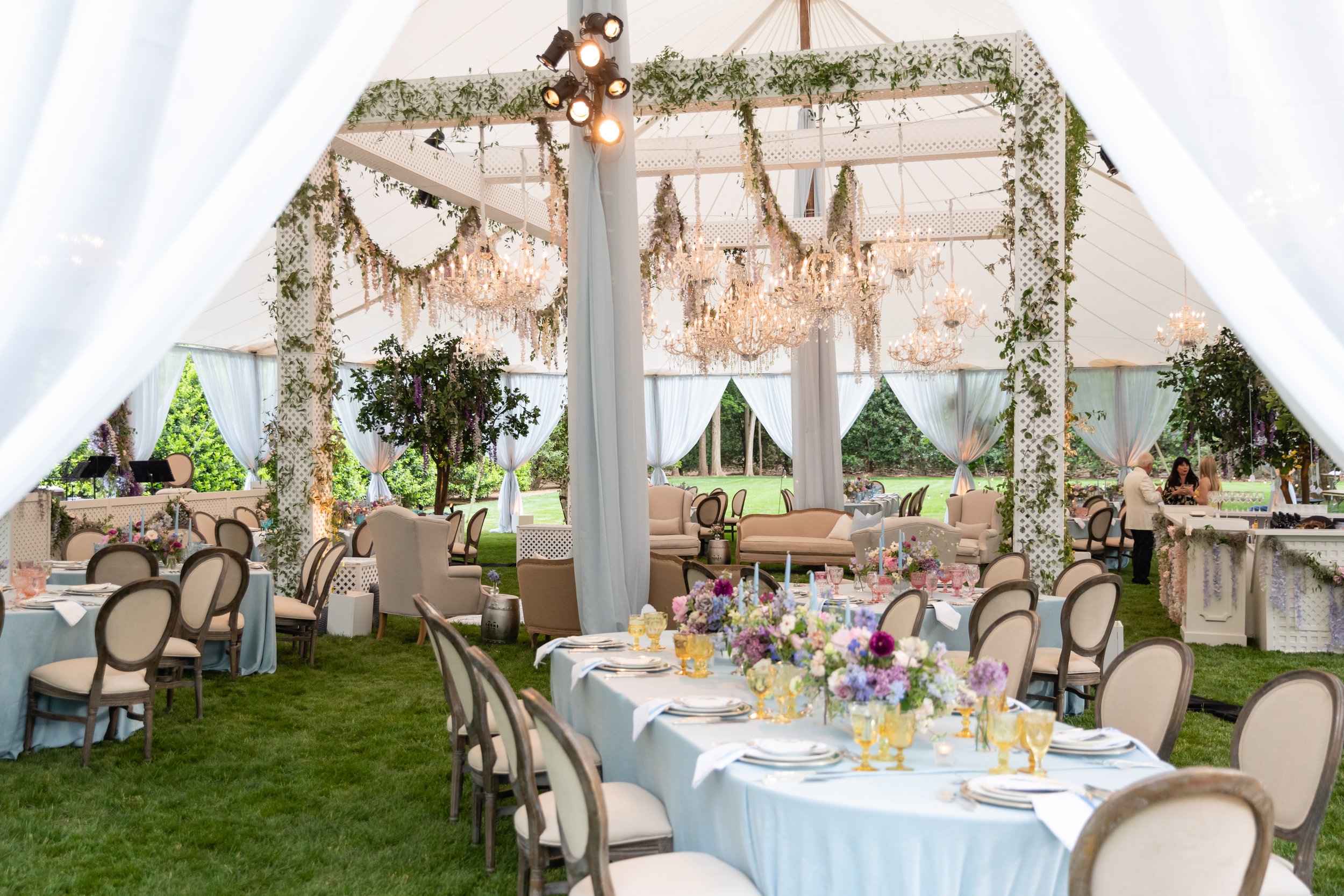 Growing, fresh floral installations and chandeliers with blush garden roses, lavender delphinium, wisteria, globe allium, and vines and greenery for a tented Bridgerton inspired engagement party at a private home in Nashville, TN.