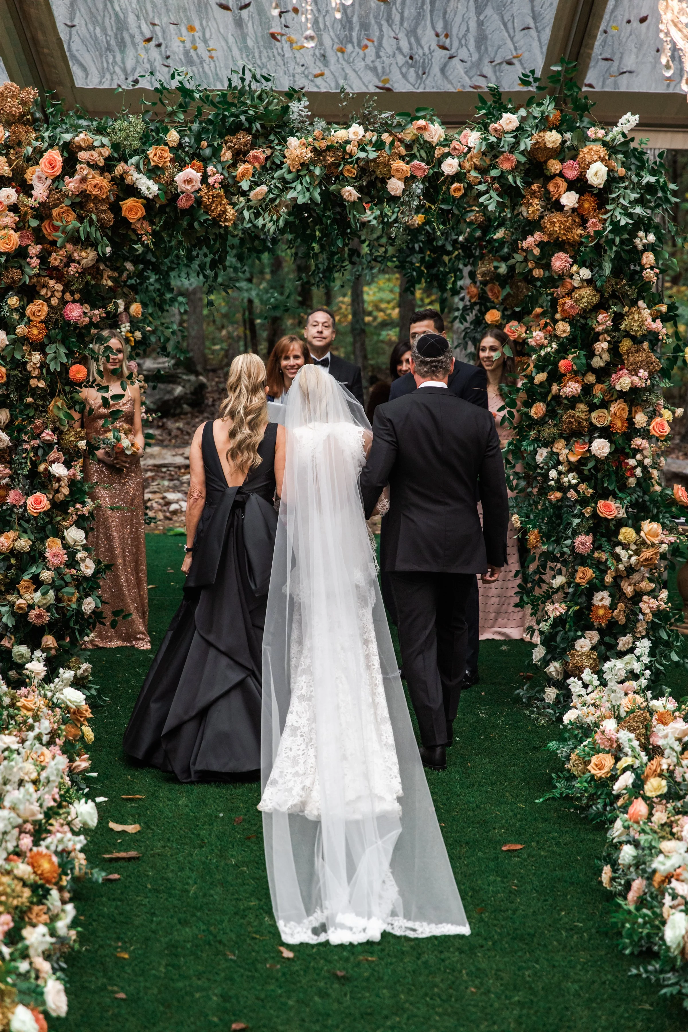 Eye-catching oversized chuppah overflowing with fall florals featuring dahlias, garden roses, rain tree pods, and fall greenery. Autumnal hues of terra cotta, dusty pink, copper, and yellow create this statement wedding piece. Designed by Rosemary an