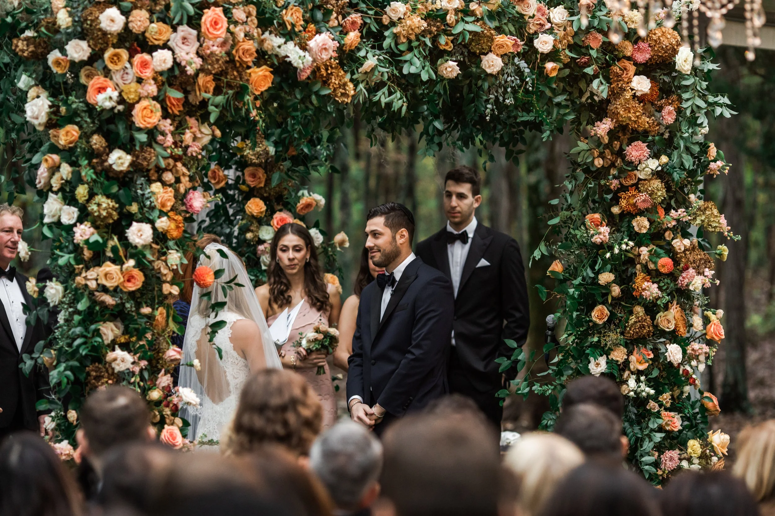 Eye-catching oversized chuppah overflowing with fall florals featuring dahlias, garden roses, rain tree pods, and fall greenery. Autumnal hues of terra cotta, dusty pink, copper, and yellow create this statement wedding piece. Designed by Rosemary an