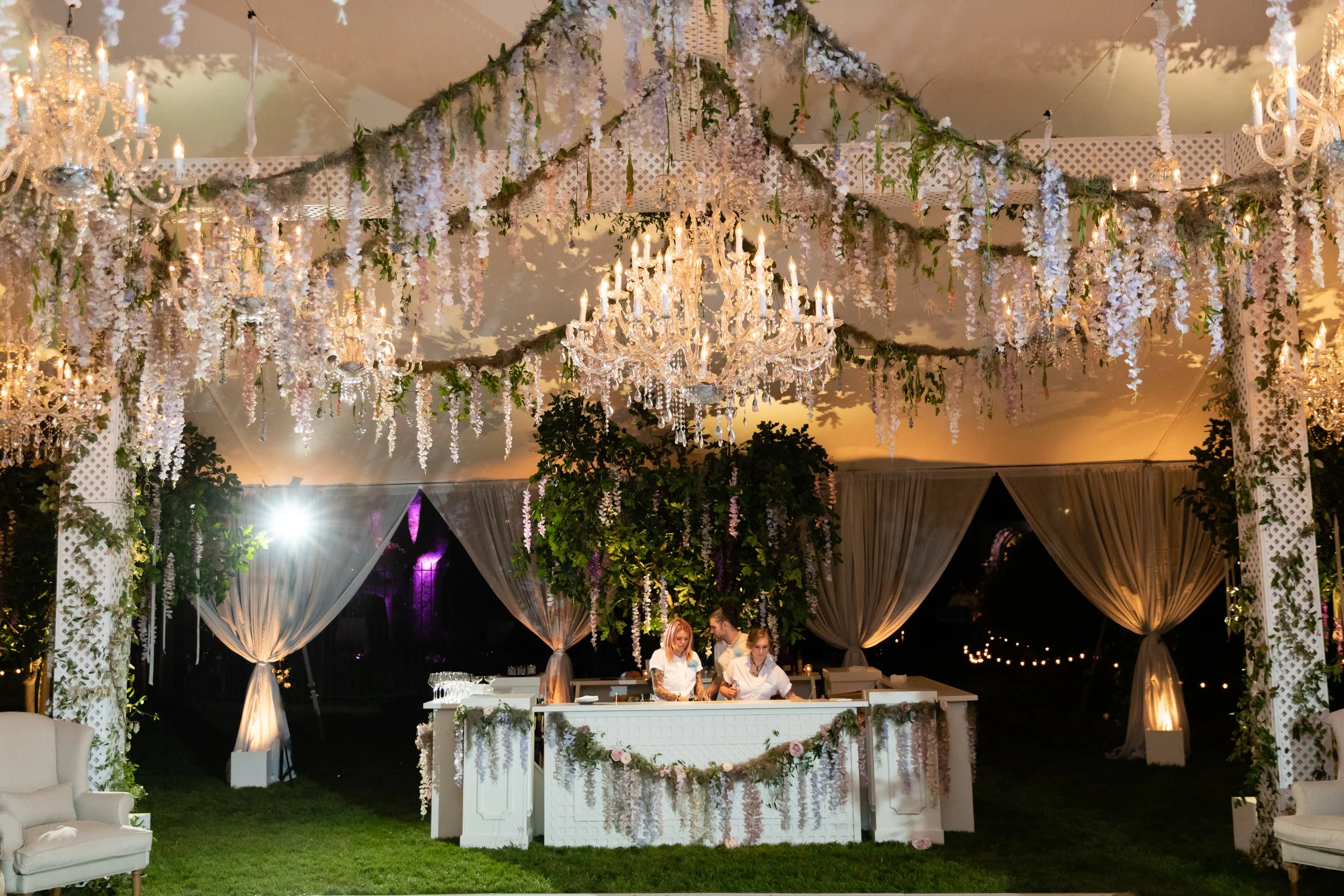 Growing, fresh floral installations and chandeliers with blush garden roses, lavender delphinium, wisteria, globe allium, and vines and greenery for a tented Bridgerton inspired engagement party at a private home in Nashville, TN.