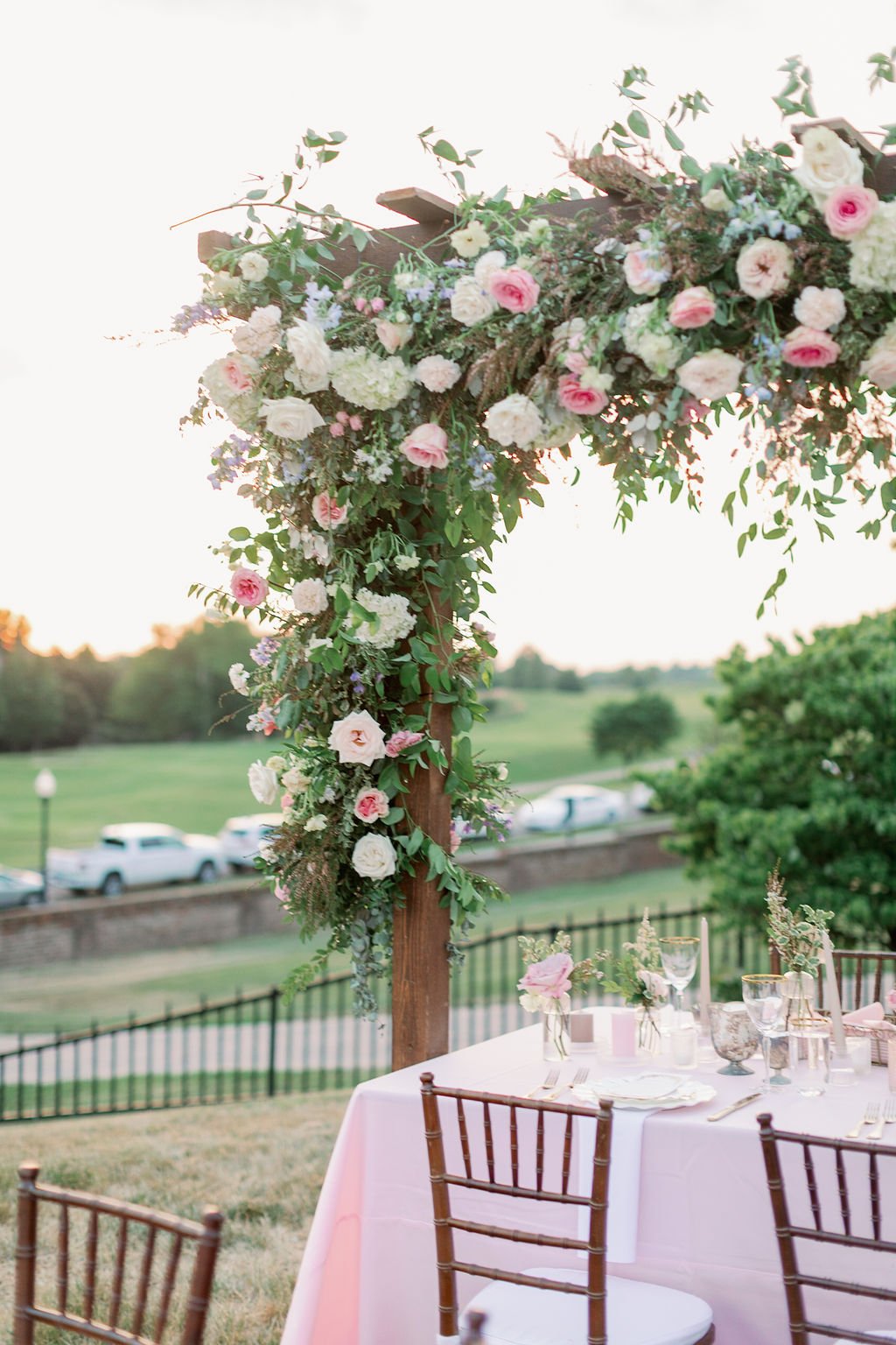 This private estate garden-inspired head table pergola is covered with pink and ivory roses, majolica spray roses, heirloom carnations, blue delphinium, hydrangea and natural greenery. Designed by Rosemary & Finch in Nashville, TN.