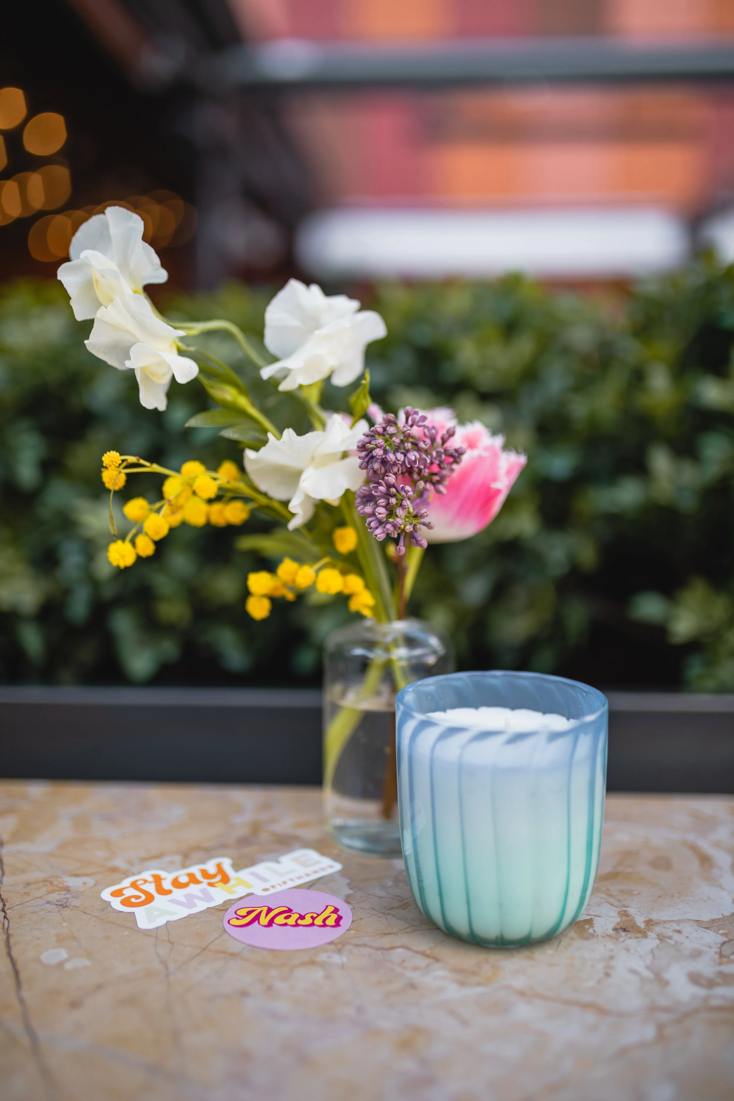 Dainty bud vases composed of roses, sweet peas, and ranunculus in floral hues of rich magenta, lavender, dusty pink, yellow, and mixed greenery. Paired with candles of unique shapes and hues. Design for Fifth + Broadway Spring by Rosemary and Finch.
