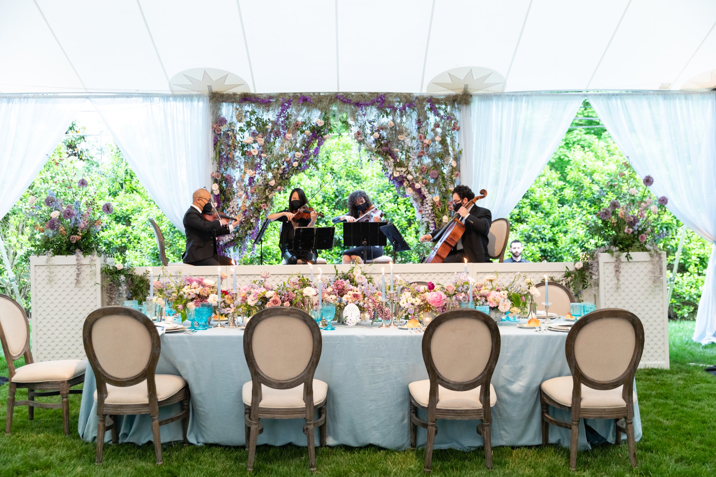 Musicians: Vitamin String Quartet. Growing, fresh floral centerpieces with a peacock, including blush garden roses, lilac, blue sweet peas, ranunculus, lavender delphinium, globe allium, and natural greenery for a Bridgerton engagement party.