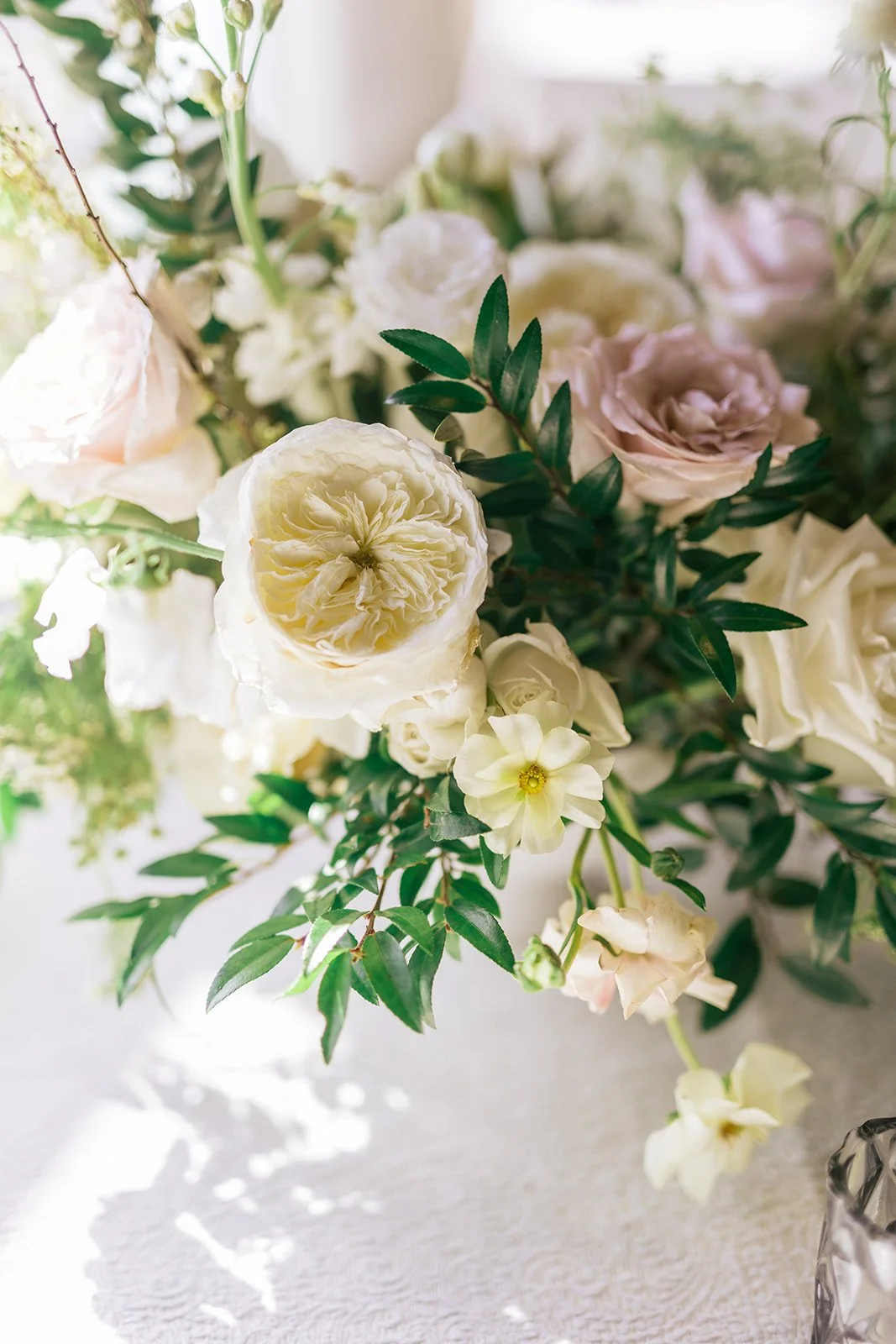 Low floral centerpieces for elegant wedding overflowing with white garden roses, ranunculus, butterfly ranunculus, scabiosa, lisianthus, sweet peas, and natural dark greenery. Floral hues of white, cream, and blush. Designed by Rosemary and Finch in
