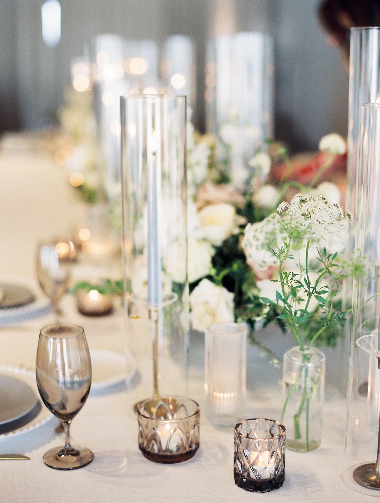 Elegant bud vases with florals of roses, ranunculus, sweet peas, Queen Anne’s lace, butterfly ranunculus in hues of white, cream and blush. Accented with dusty blue tapers and clear glass votives. Designed by Rosemary and Finch in Nashville, TN.