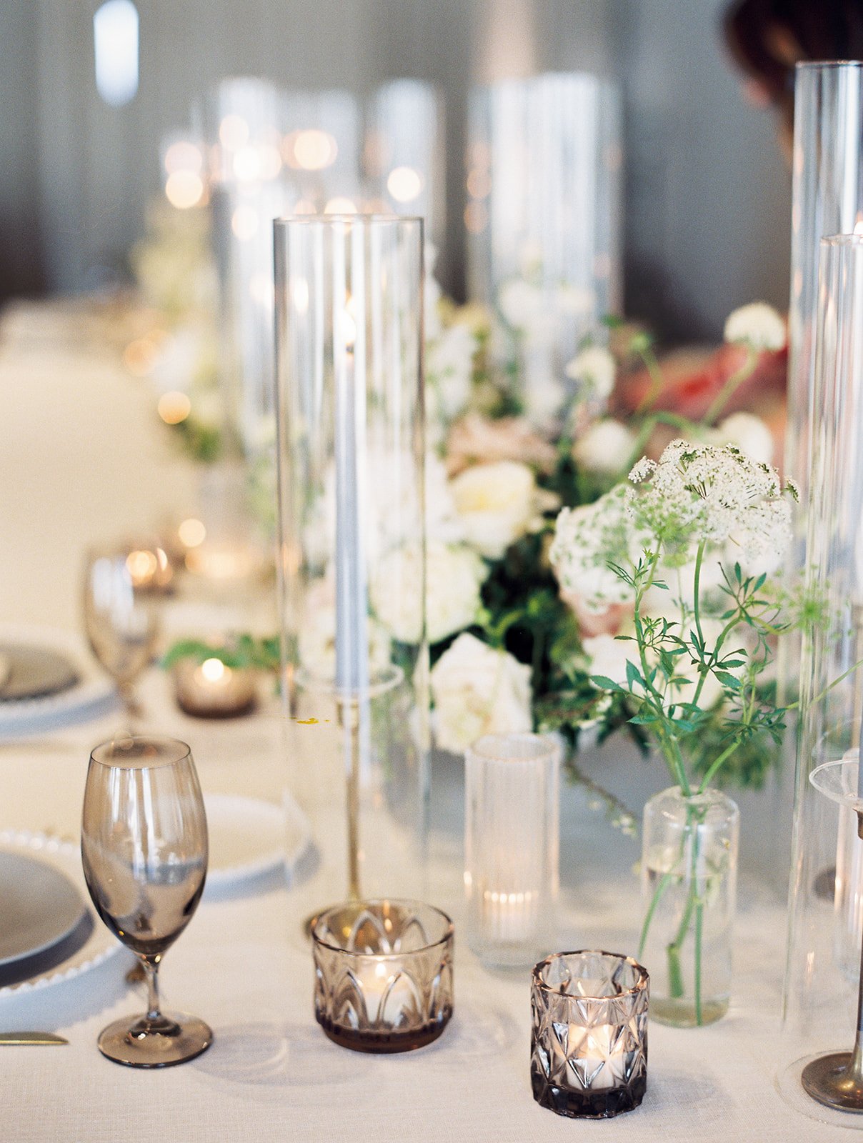 Elegant bud vases with florals of roses, ranunculus, sweet peas, Queen Anne’s lace, butterfly ranunculus in hues of white, cream and blush. Accented with dusty blue tapers and clear glass votives. Designed by Rosemary and Finch in Nashville, TN.