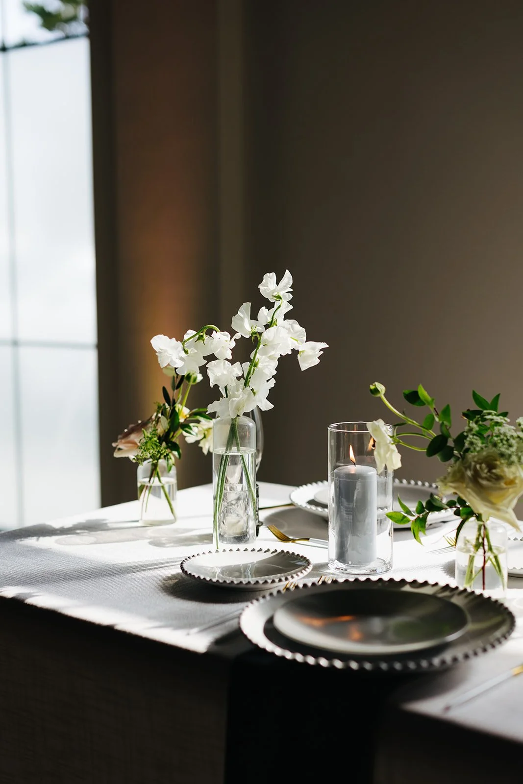 Elegant bud vases with florals of roses, ranunculus, sweet peas, Queen Anne’s lace, butterfly ranunculus in hues of white, cream and blush. Accented with dusty blue tapers and clear glass votives. Designed by Rosemary and Finch in Nashville, TN.