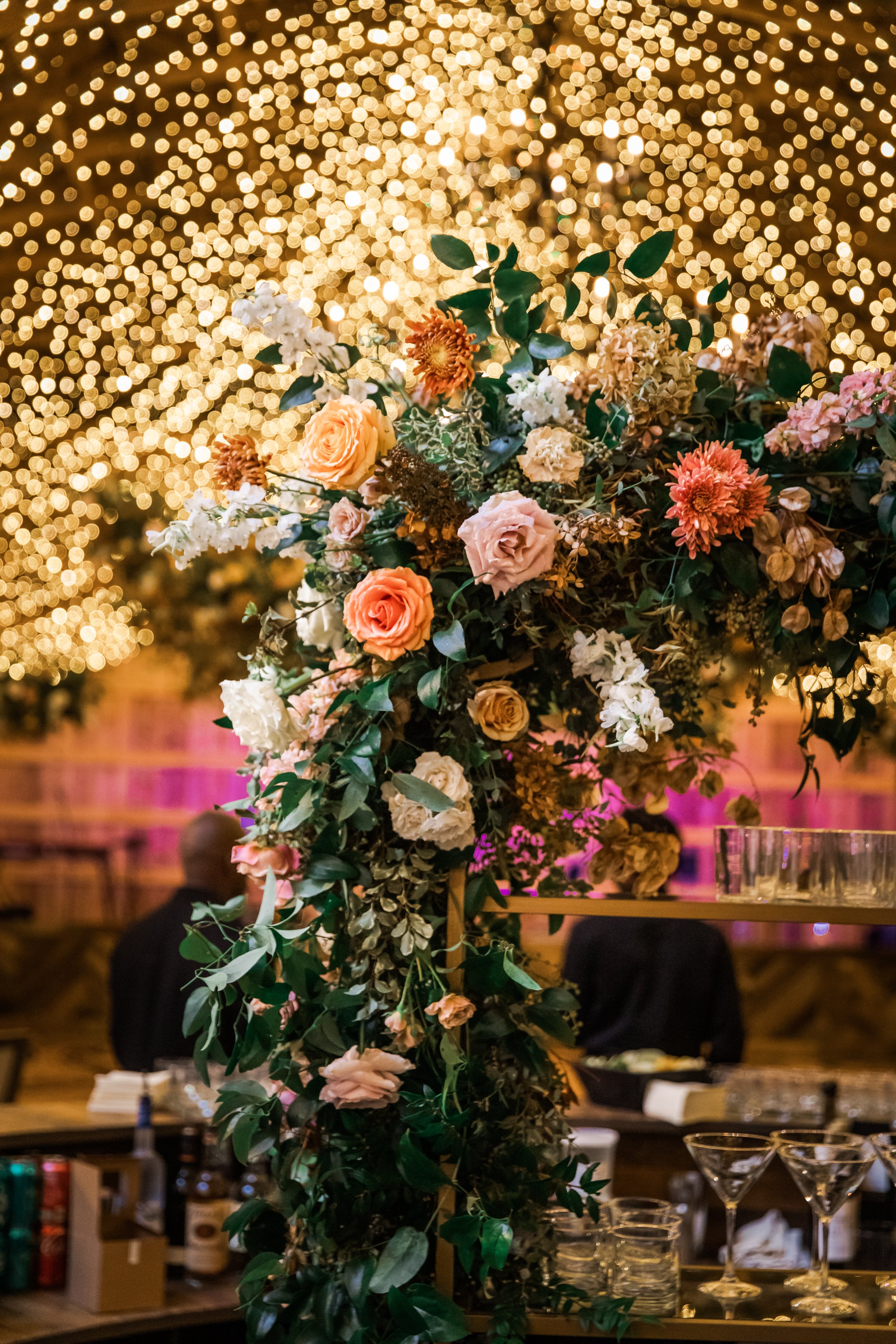 Stunning fall floral installation frame the bar area of this autumnal wedding. Hues of blush, terra cotta, copper, toffee, yellow, and other neutrals are highlighted with dahlias, roses, and double brownie tulips. Designed by Rosemary and Finch in Na