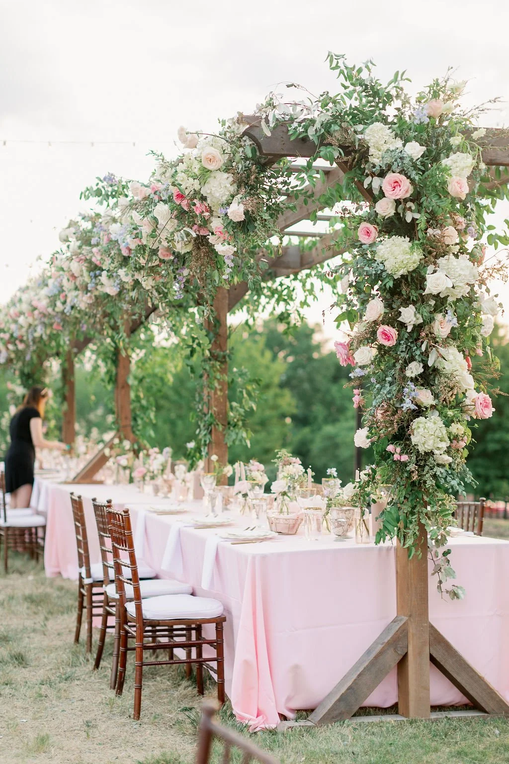 This private estate garden-inspired head table pergola is covered with pink and ivory roses, majolica spray roses, heirloom carnations, blue delphinium, hydrangea and natural greenery. Designed by Rosemary & Finch in Nashville, TN.