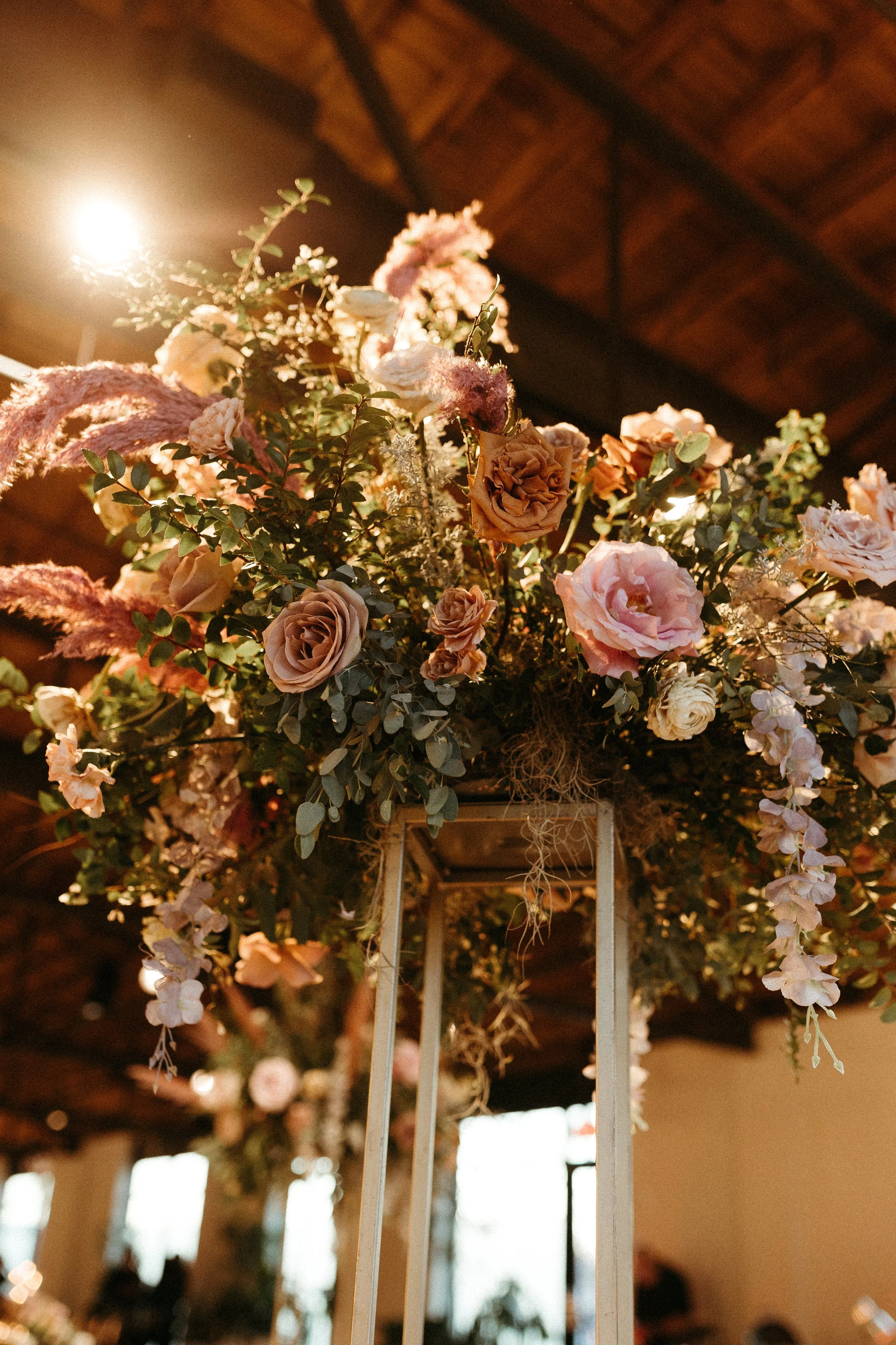 Stunning elevated centerpieces decorate this art deco wedding. Floral hues of terra cotta, mauve, burgundy, and dusty pink are brought to life with petal heavy roses, dried branches, and pink pampas grass. Designed by Rosemary and Finch in Nashville,
