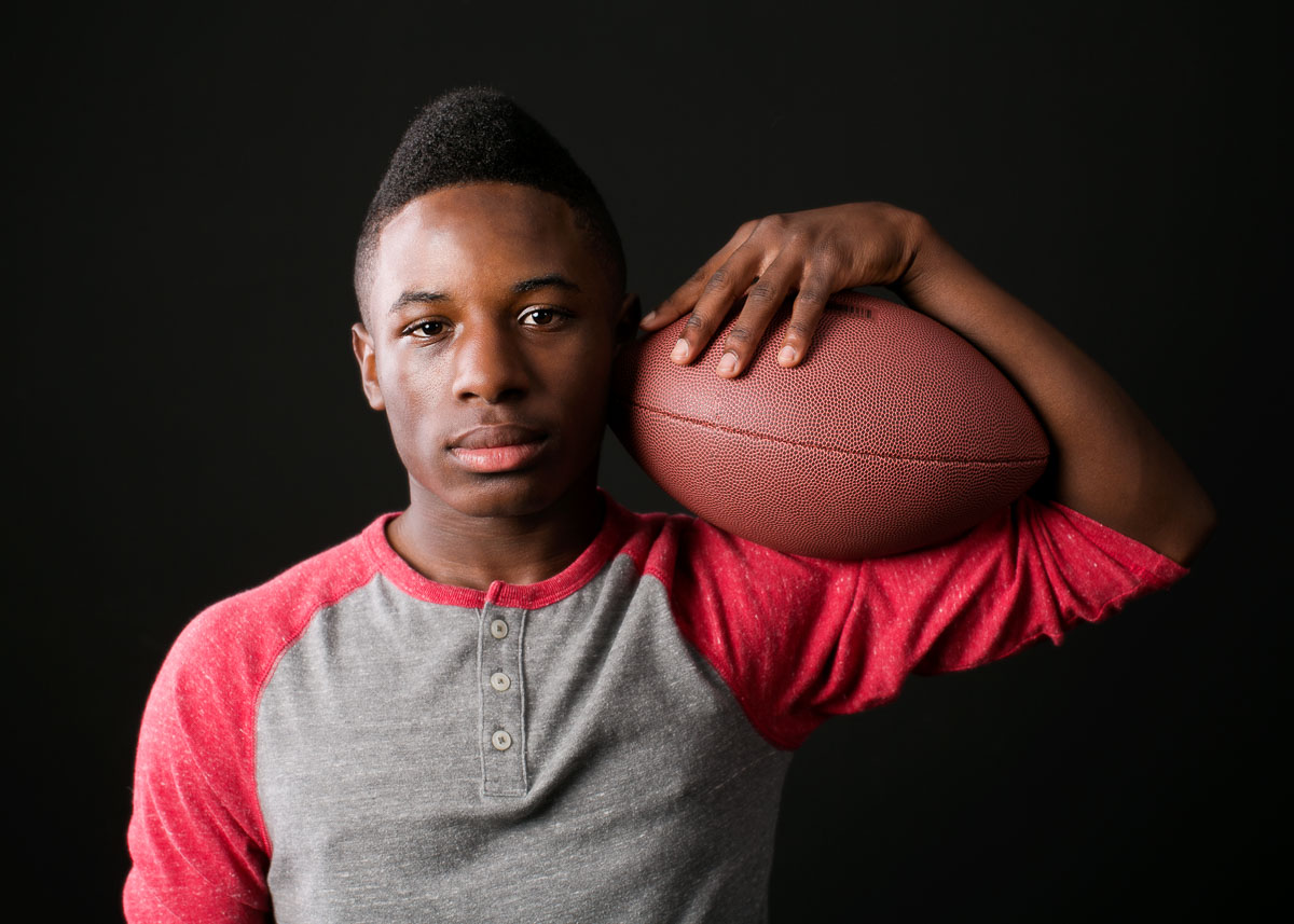  High school senior photography of a young man holding a football against a black background by Campbell Salgado Studio. 