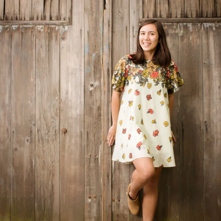 Young woman in front of barn doors in a outdoor Campbell Salgado Studio for her high school senior pictures session. Wearing gray and purple against green foliage.