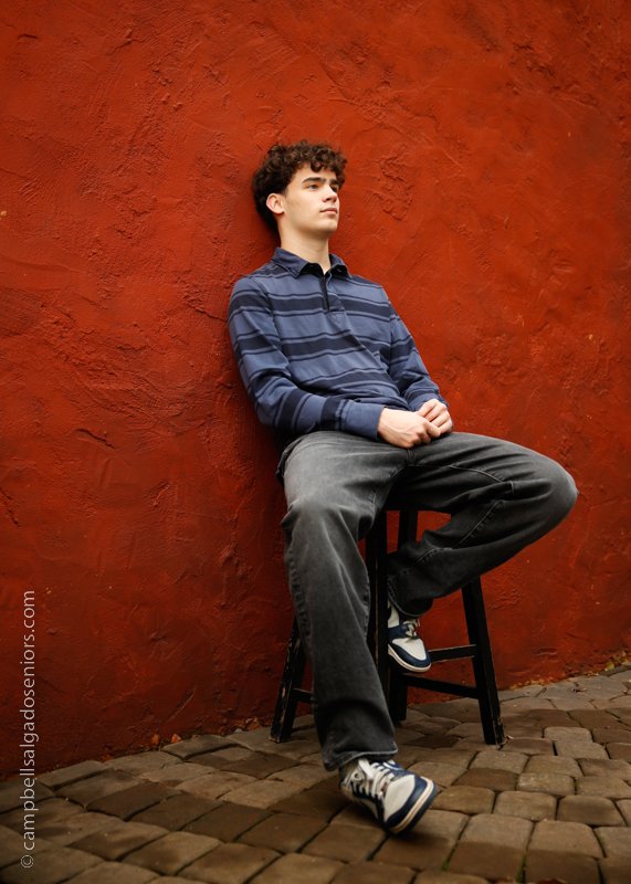  High school senior pictures&nbsp;of a young man standing in front of&nbsp;red stucco background by senior photographers at Campbell Salgado Studio in Portland, Oregon. 