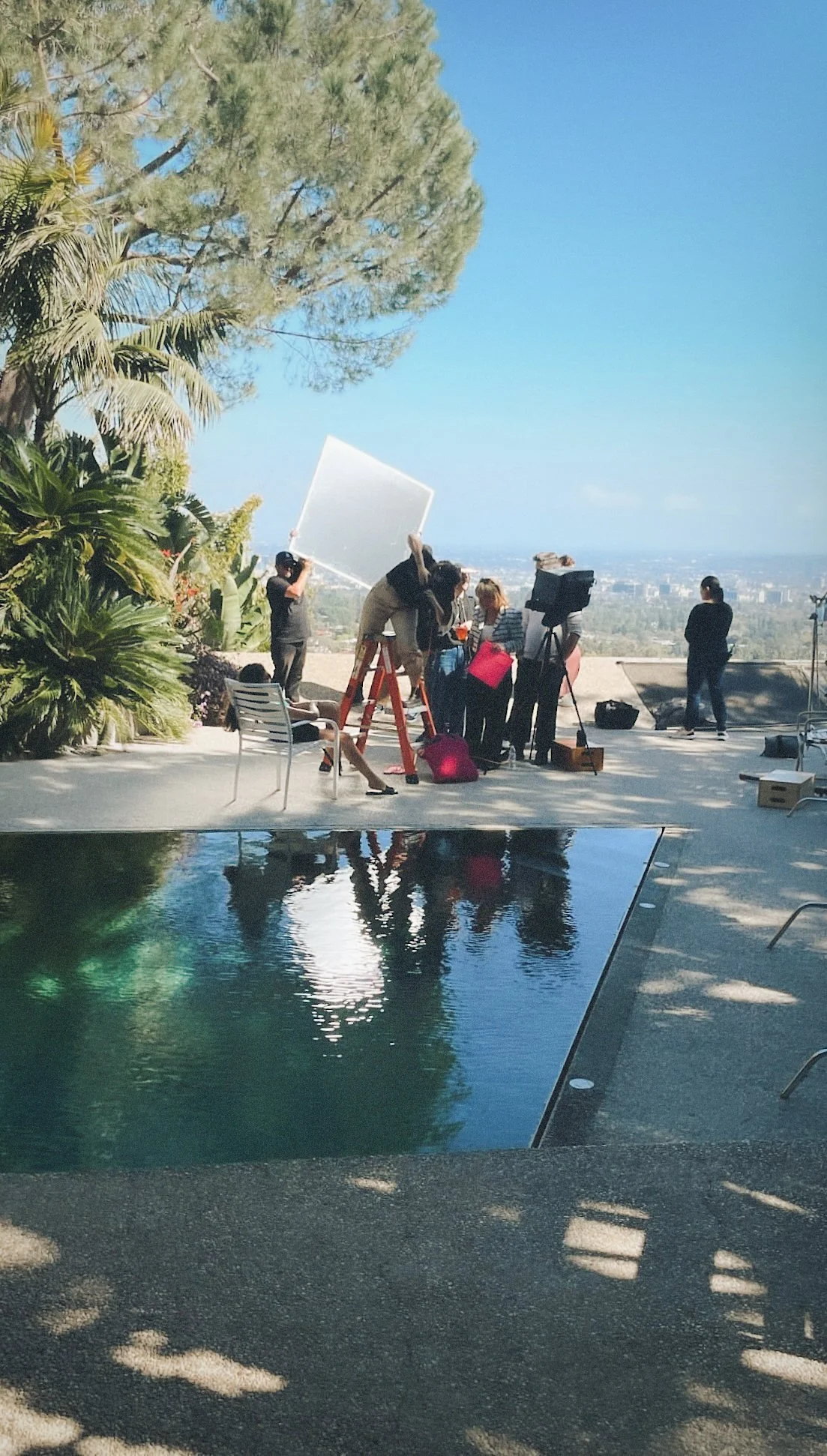 Film crew filming a scene outdoors near a swimming pool with crew members setting up equipment and a director.