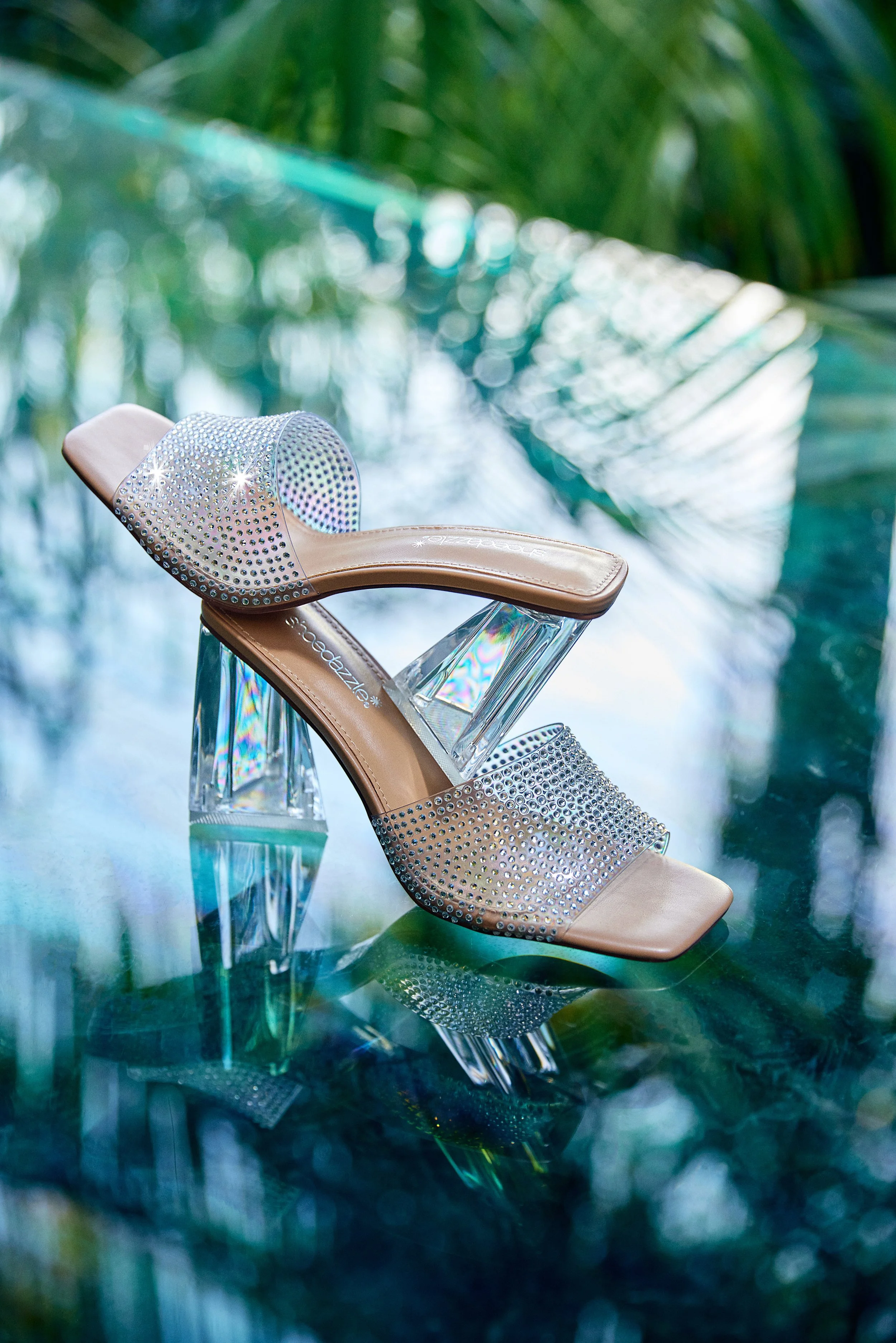 A high-heeled sandal with rhinestones, a clear, geometric heel, and a beige insole, displayed on a reflective surface against a blurred background of palm leaves and sunlight.