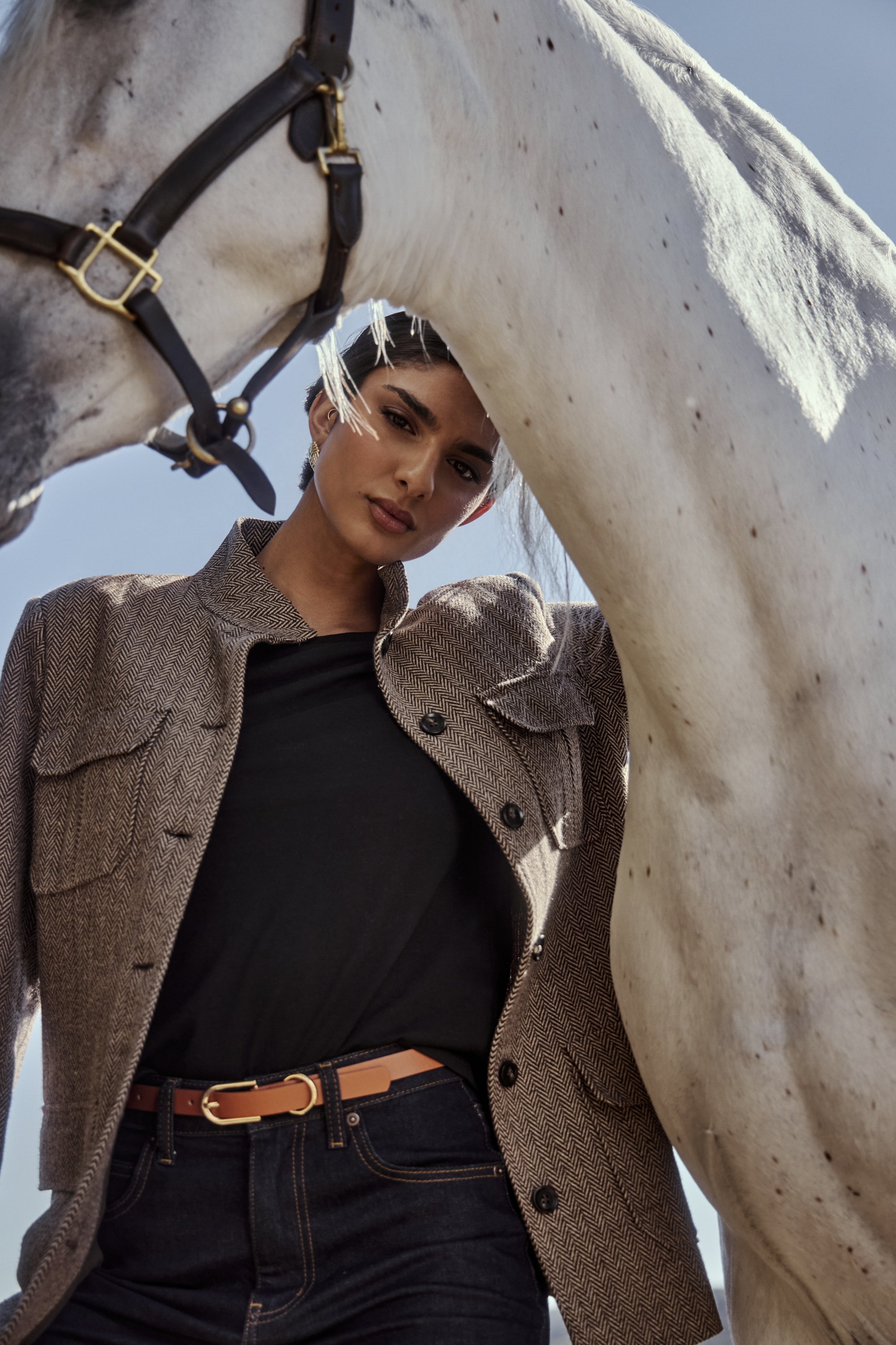 A woman standing next to a white horse, looking down at the camera, with the horse's head in the foreground, against a clear blue sky.