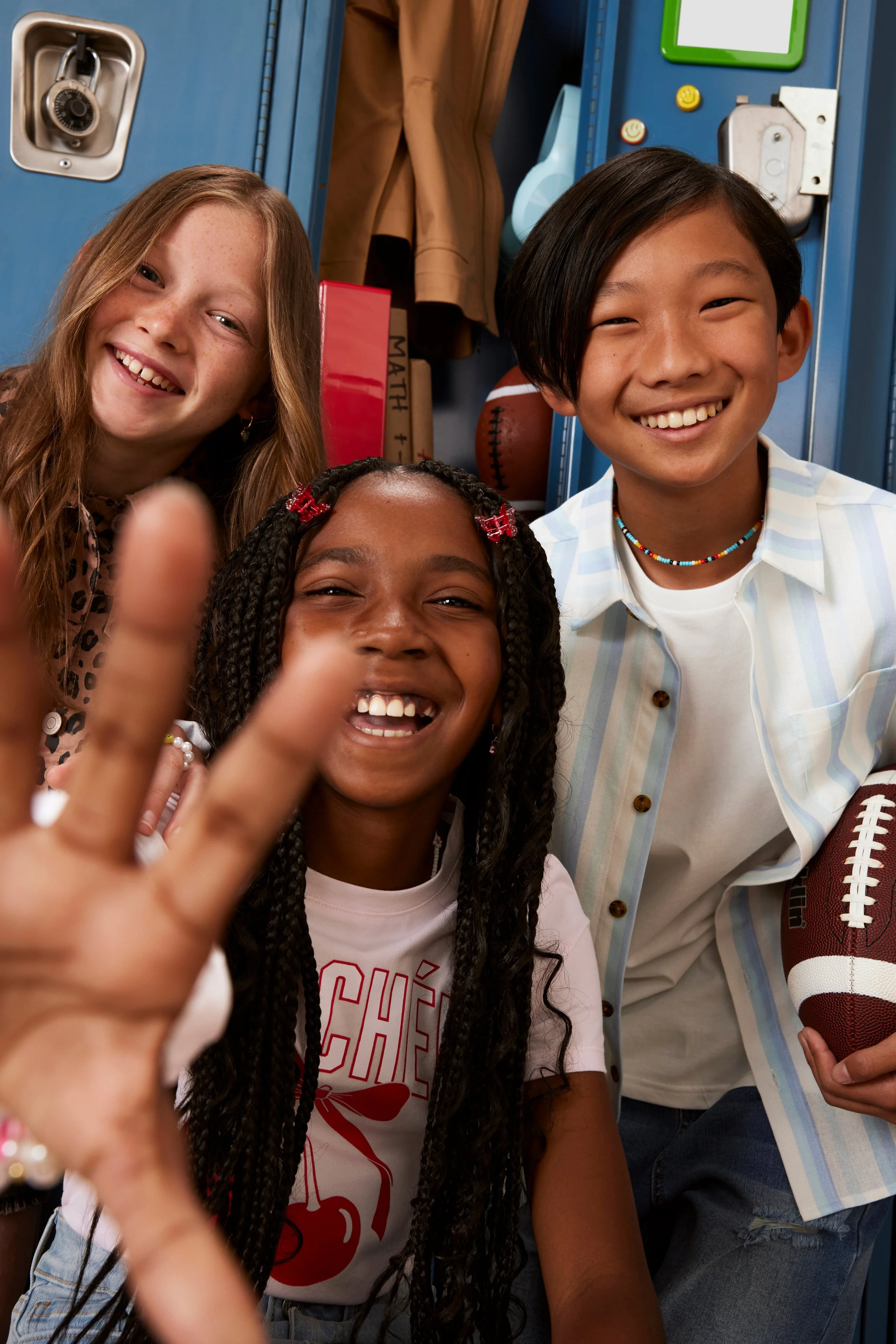 Three smiling students inside a school locker room, with one holding a football, and locker items in the background.