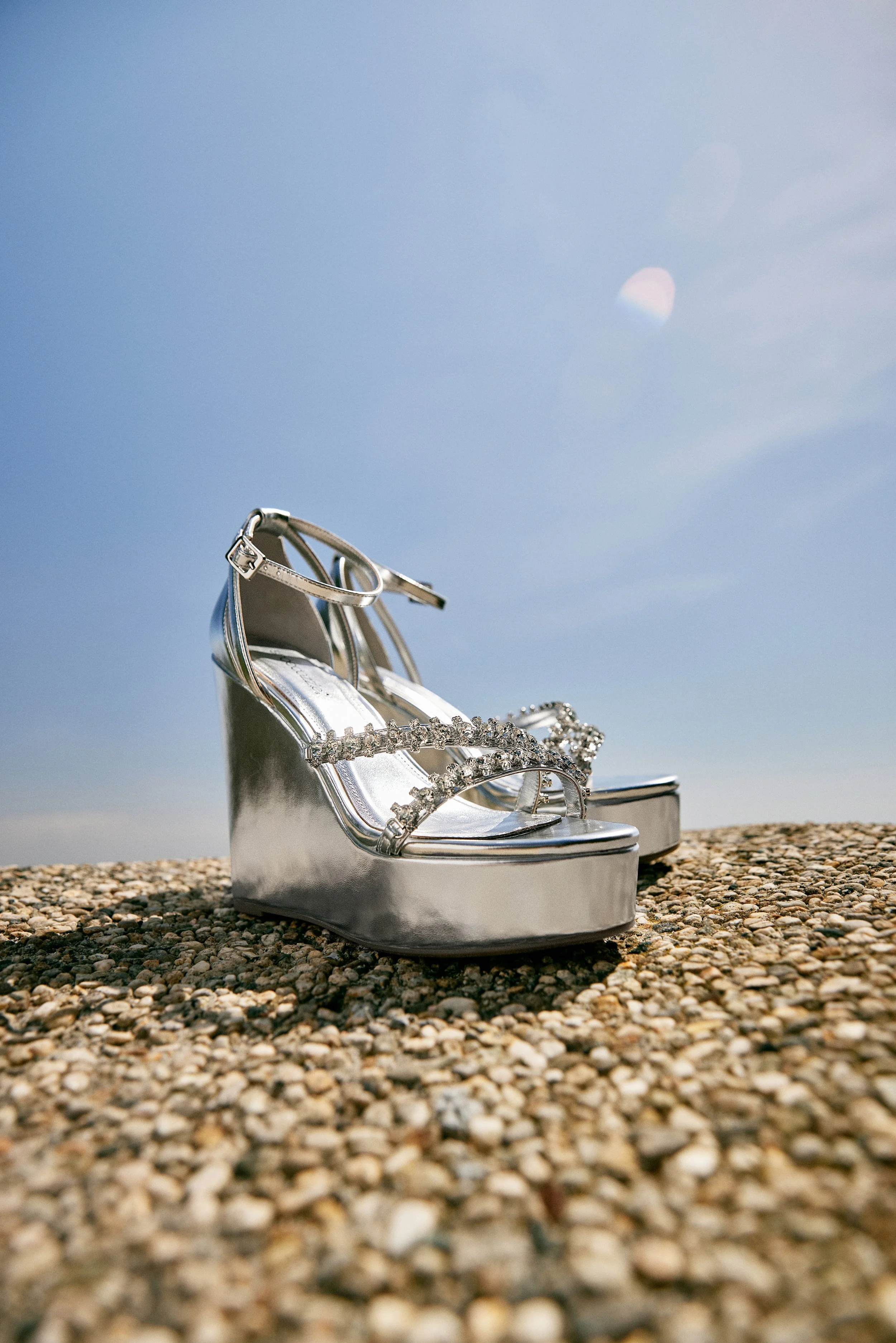 Silver high heel shoes with rhinestone straps on rocky ground against a blue sky.