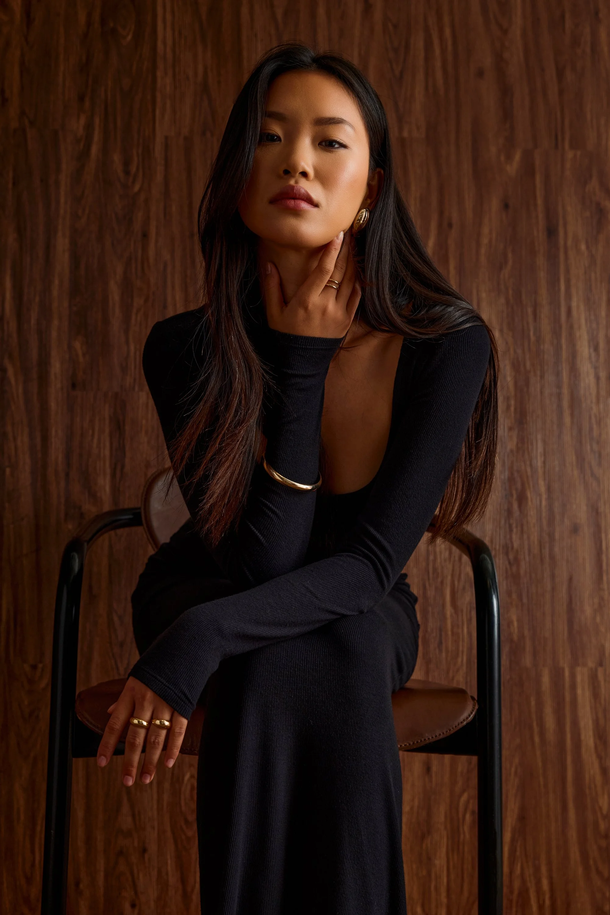 A woman with long dark hair, wearing a black long-sleeve top and black pants, sits on a chair against a wooden background. She is touching her neck with her right hand and resting her left arm on her leg, wearing gold jewelry.