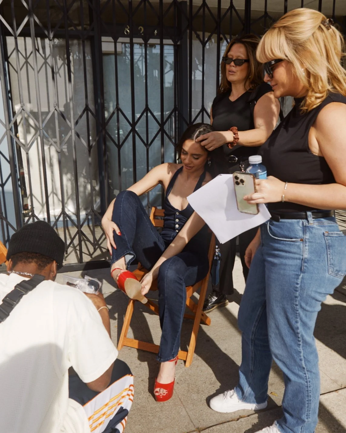 A woman sitting in a director's chair getting her foot checked by a person kneeling on the ground, surrounded by three other women standing nearby, one holding a smartphone and a water bottle, outside near a metal fence.
