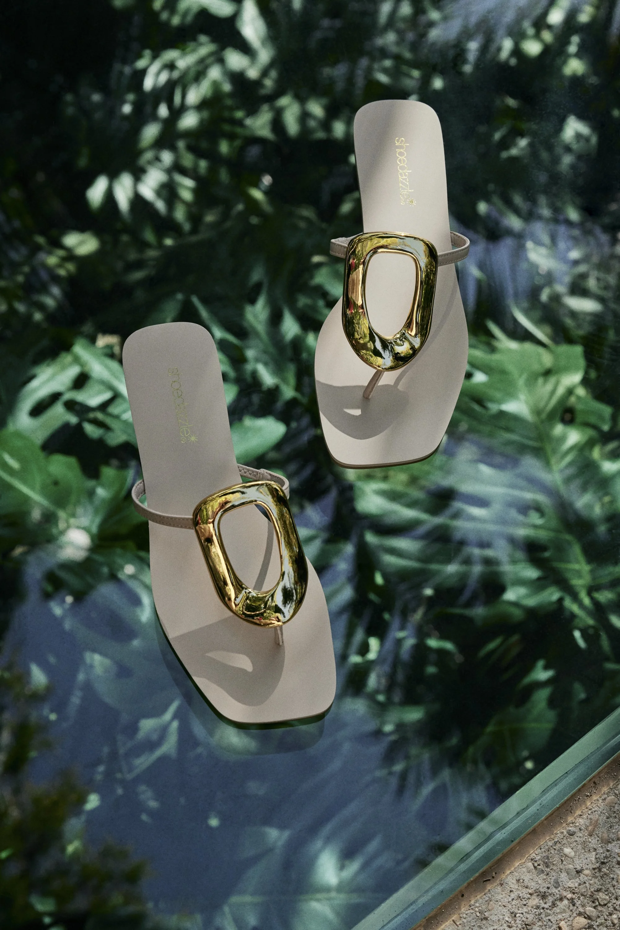 Pair of beige sandals with large gold buckle embellishments, displayed on a glass surface, with lush green leaves in the background.