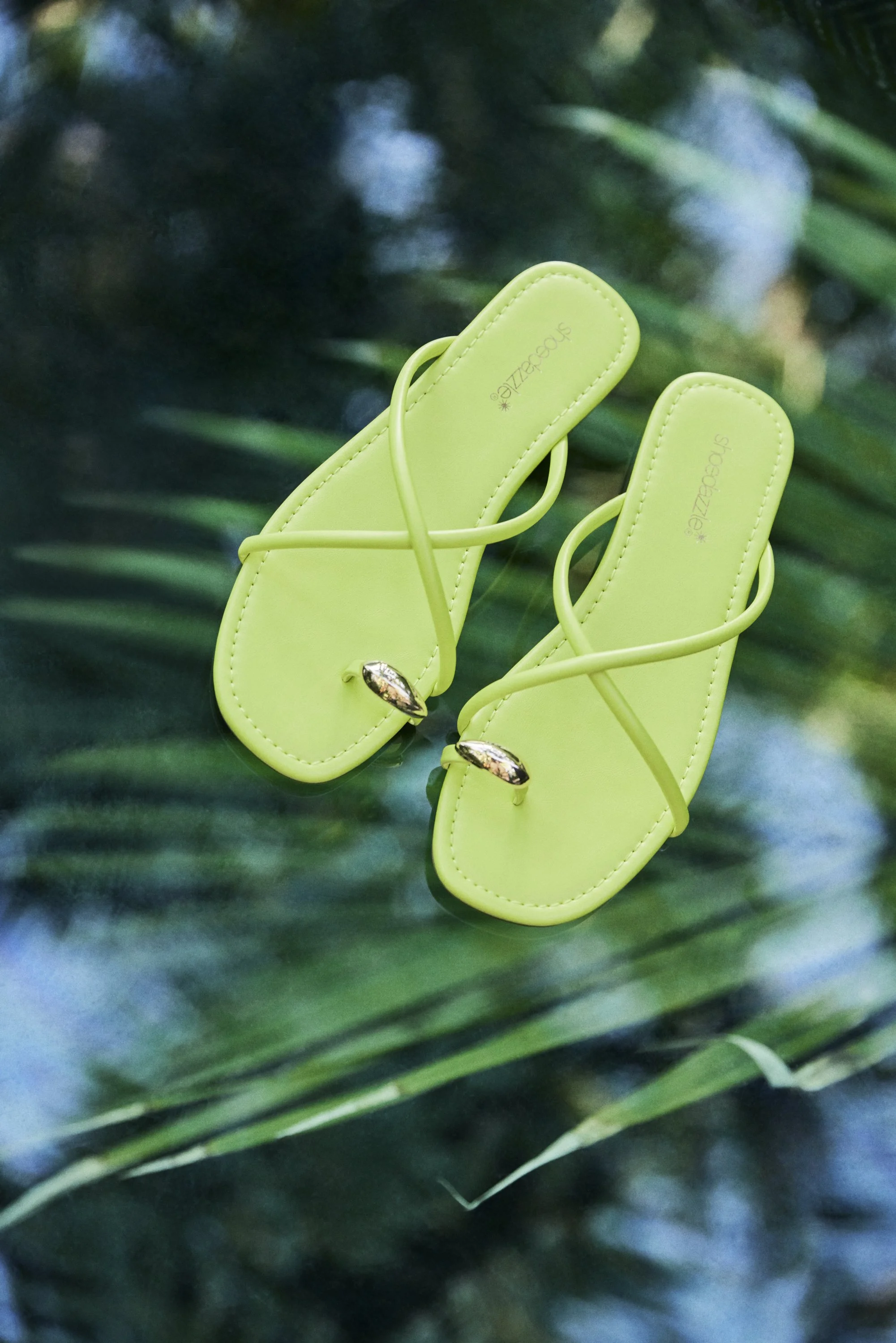 Bright yellow-green sandals with thin straps and small decorative heels resting on green leaves.