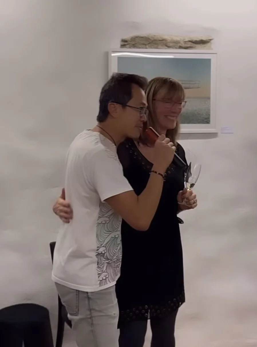 Two people hugging at an art gallery, one holding a glass of wine and the other using a mobile phone, with a framed artwork on the wall behind them.