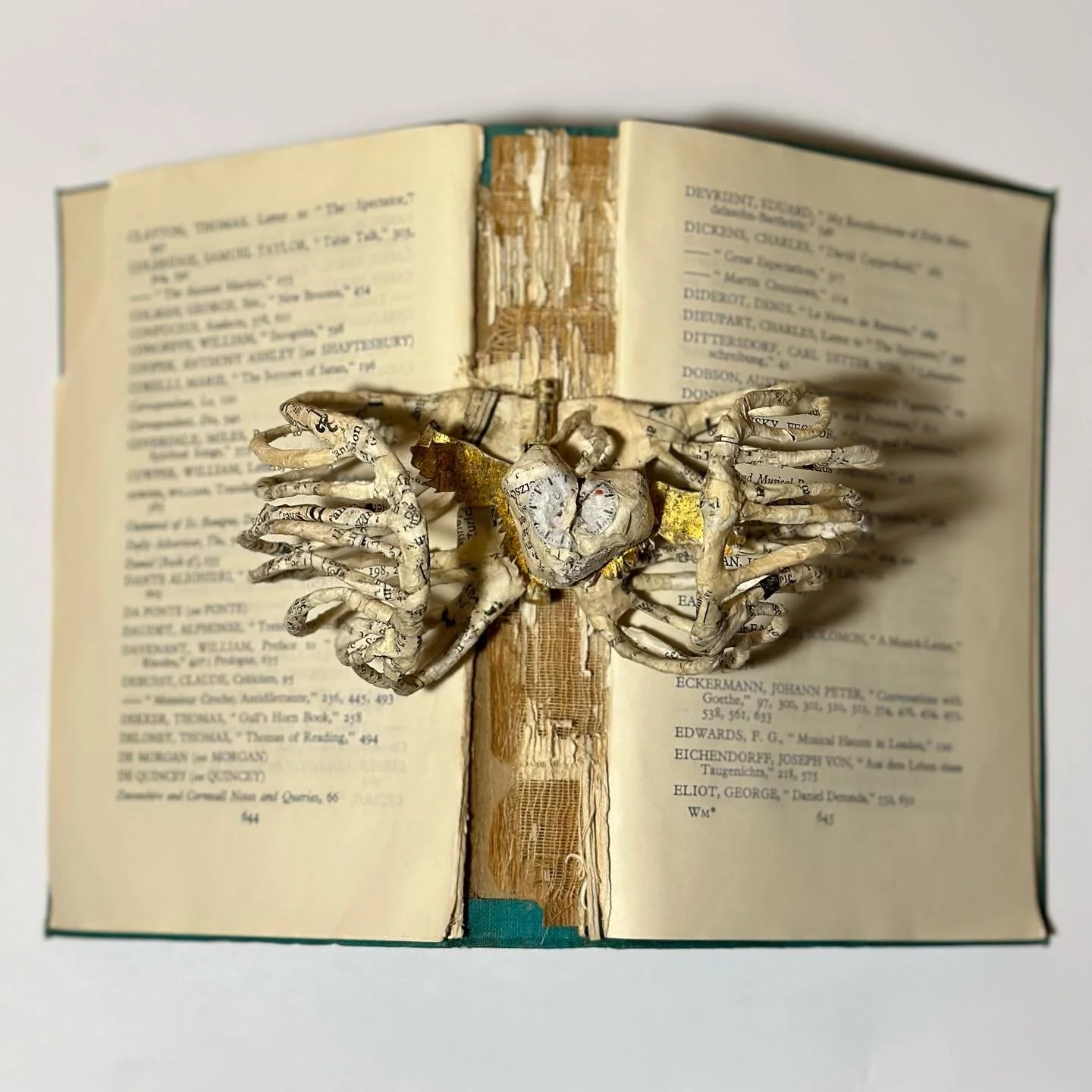 A small sculpture of a ribcage made from vintage newspaper pages in papier mache, mounted on an open book with added gold accents.