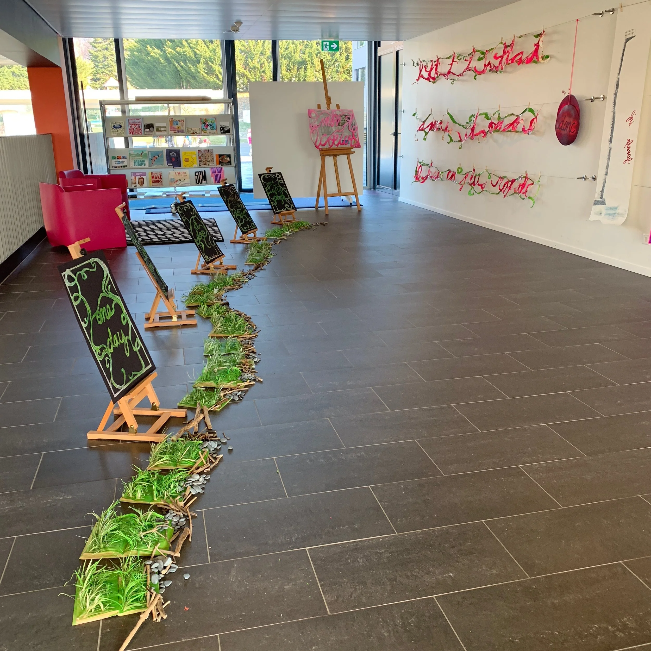 Indoor space with black tiled floor and large windows. Decorative display includes easels and papercuts with green decorative writing. There is a white rotating bookshelf filled with books, pink armchairs, and more papercut writing.
