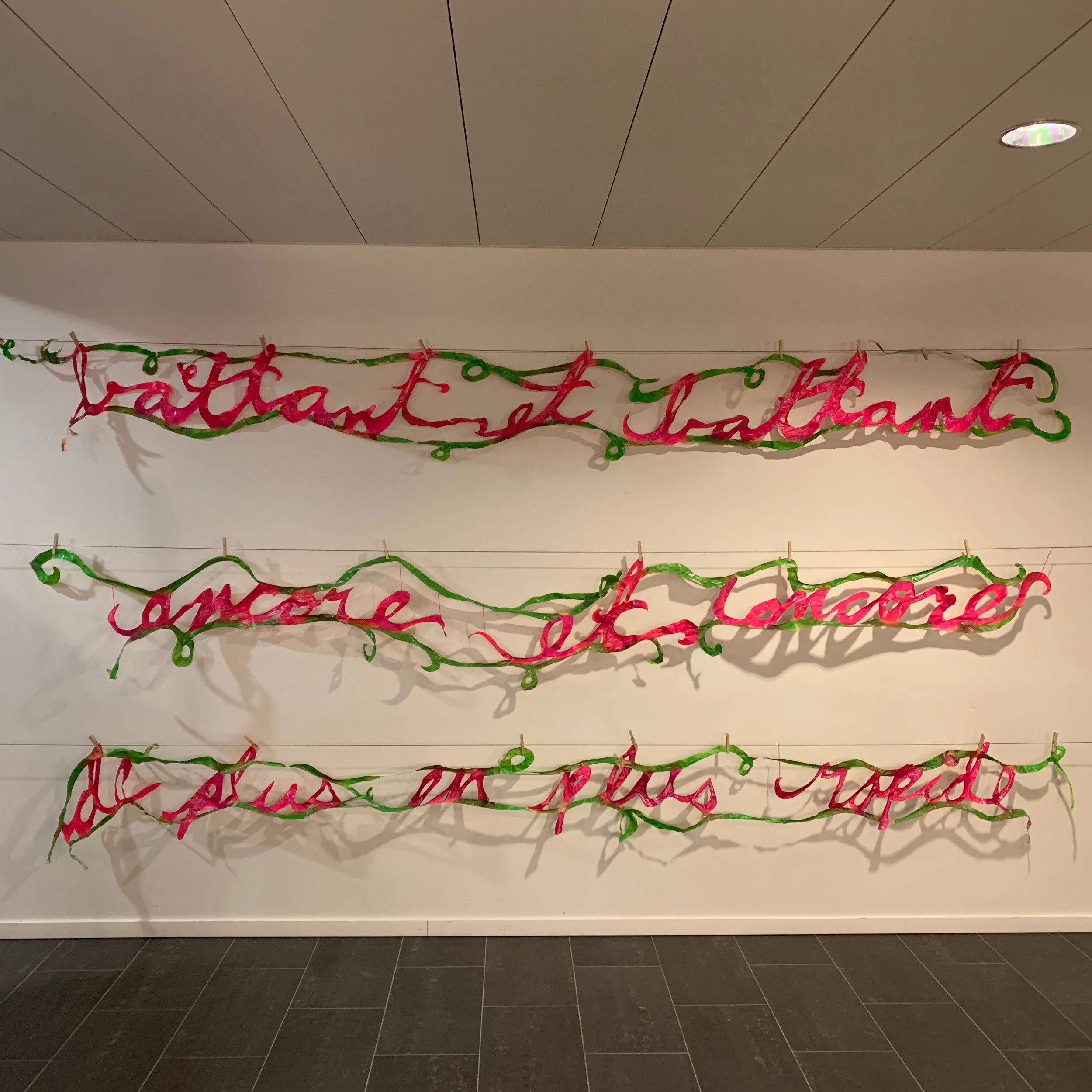 Decorative papercut banners with pink and green text that says battant et battant, encore et encore, plus en plus rapide' hanging on a white wall with horizontal lines.