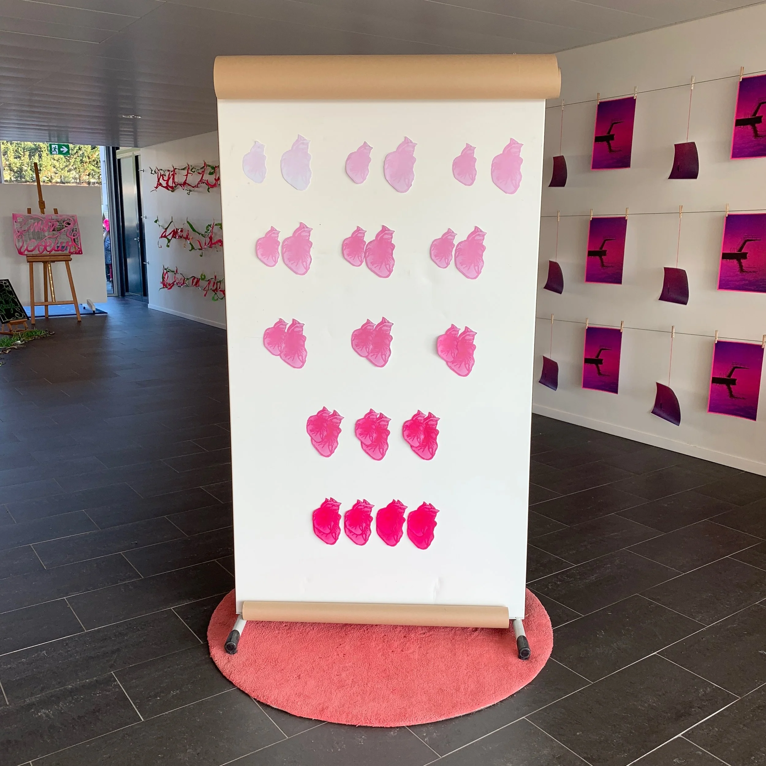 Pink gradient heart prints display board in an art gallery, surrounded by other artwork including hanging photos and hanging text elements.
