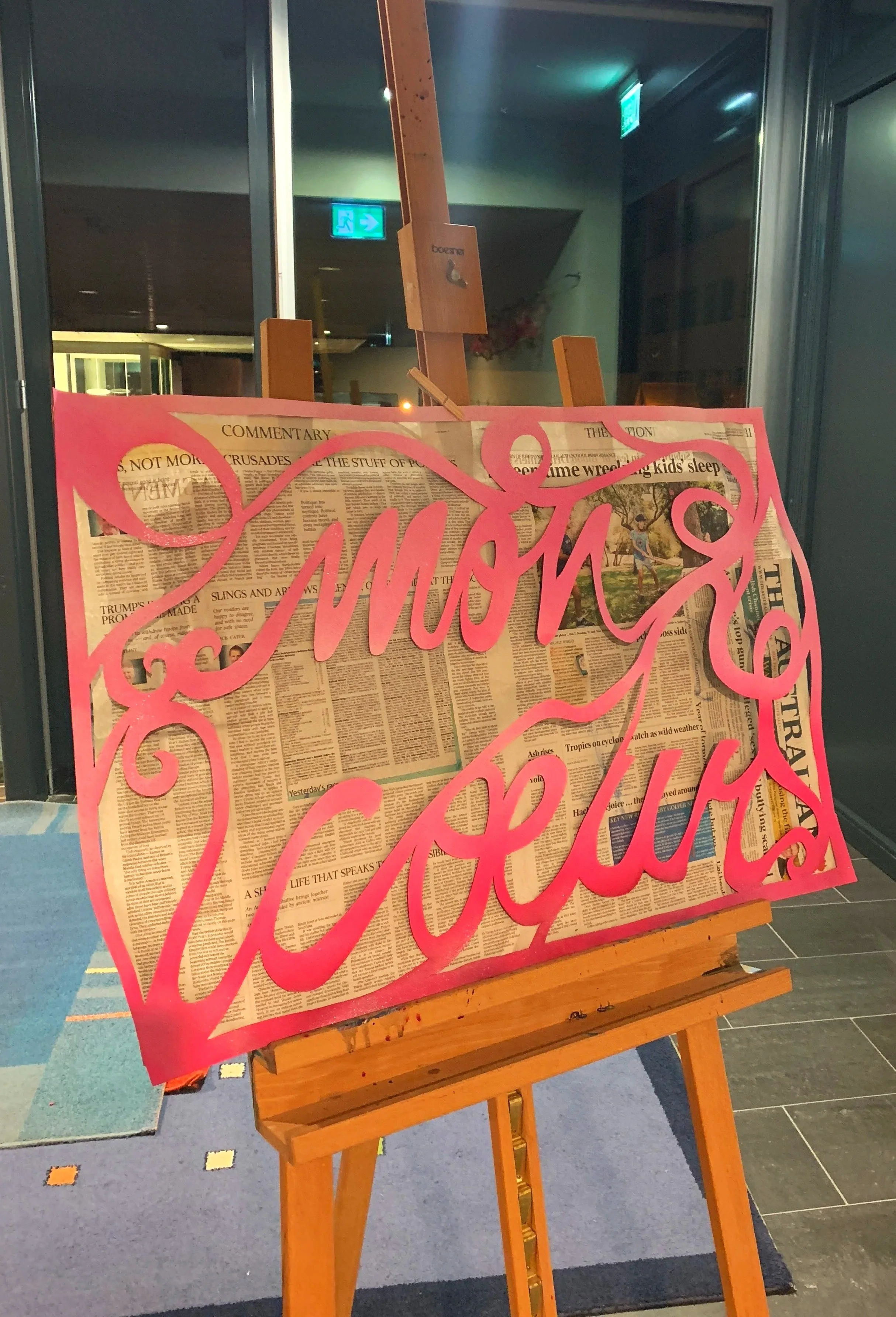 A wooden easel holding a large piece of newspaper covered with newspaper and pink cursive writing. The background includes glass doors, a lit exit sign, and a dark interior space.
