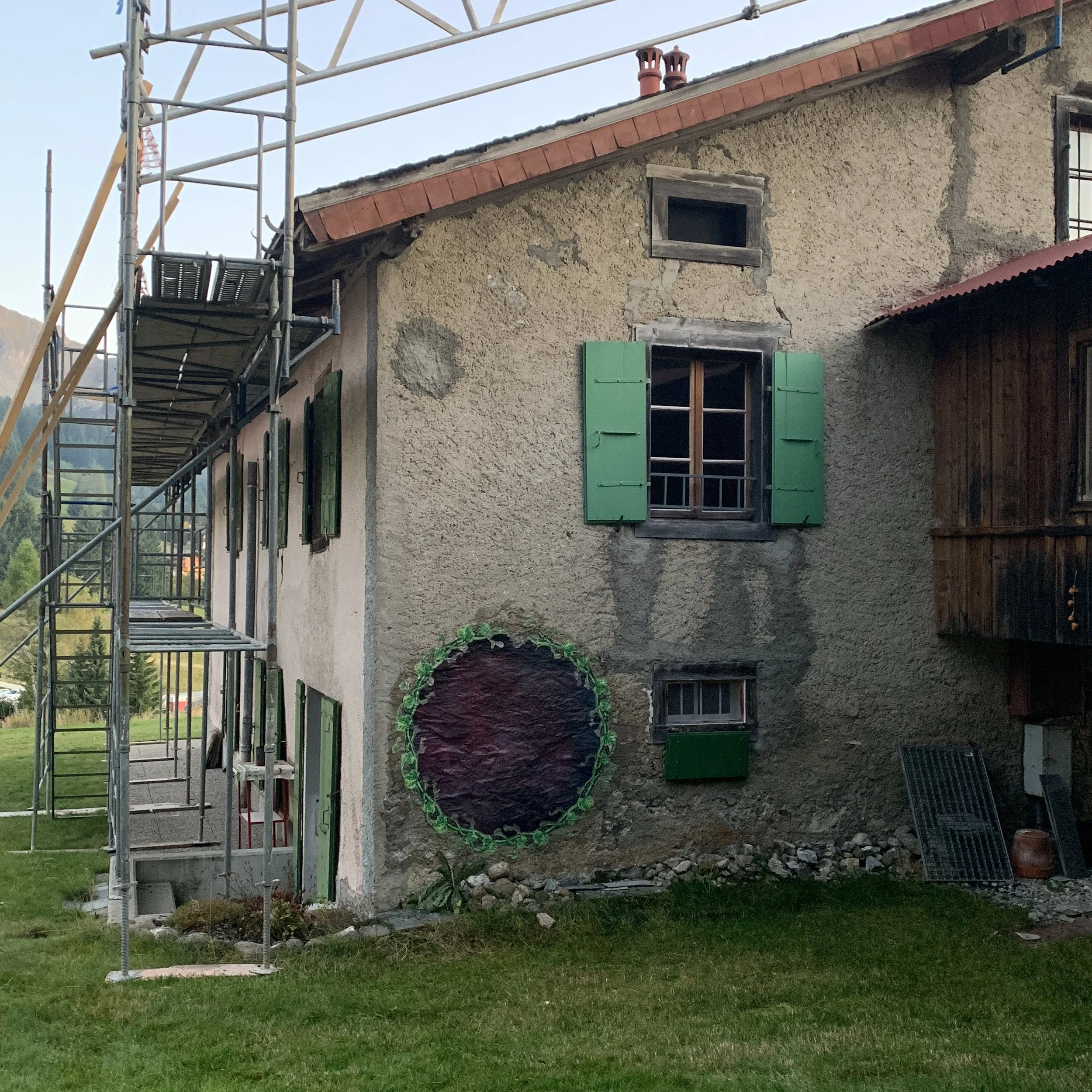 A house under renovation with scaffolding on the left side. The house has a beige stucco exterior with visible cracks and two small windows with green shutters. The roof has red tiles, and there are two red chimney pipes on top. There is a creatively decorated round object, possibly a Halloween decoration, on the bottom left.