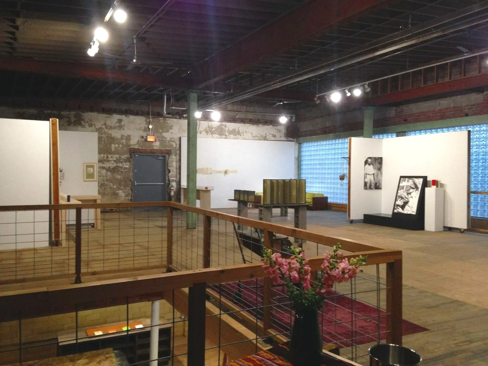 Interior of an industrial-style building with exposed brick and metal ceiling, 3546 Michigan Ave Detoit Holding House, spotlights, white display boards, and art. A staircase with a wooden handrail and planter with pink flowers are visible.