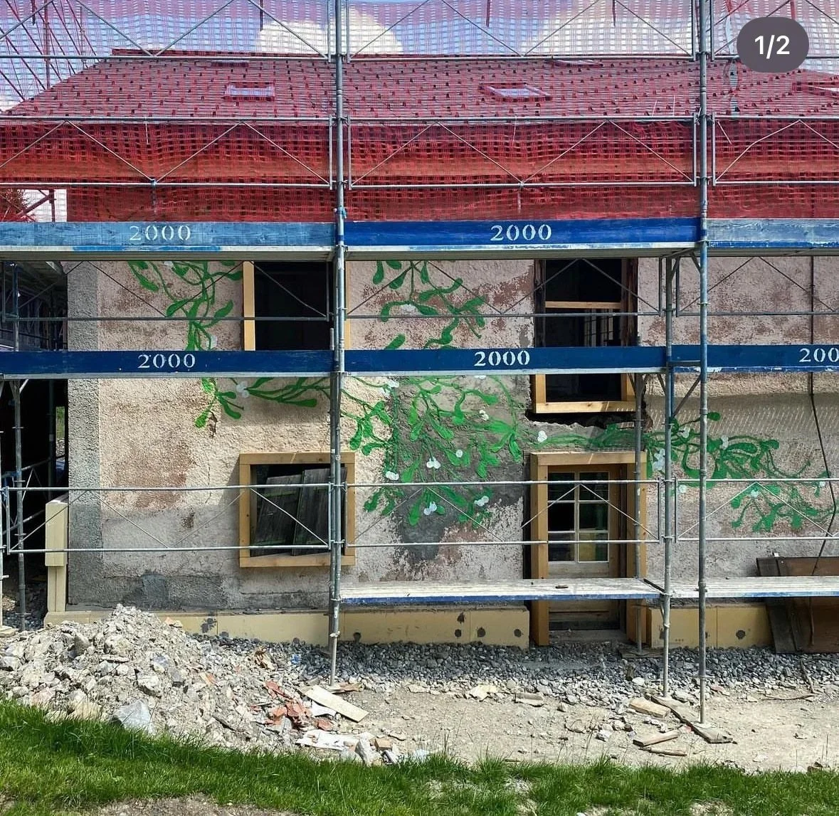 A building under construction with scaffolding in front, showing a mural of green tree branches painted on an exterior wall.